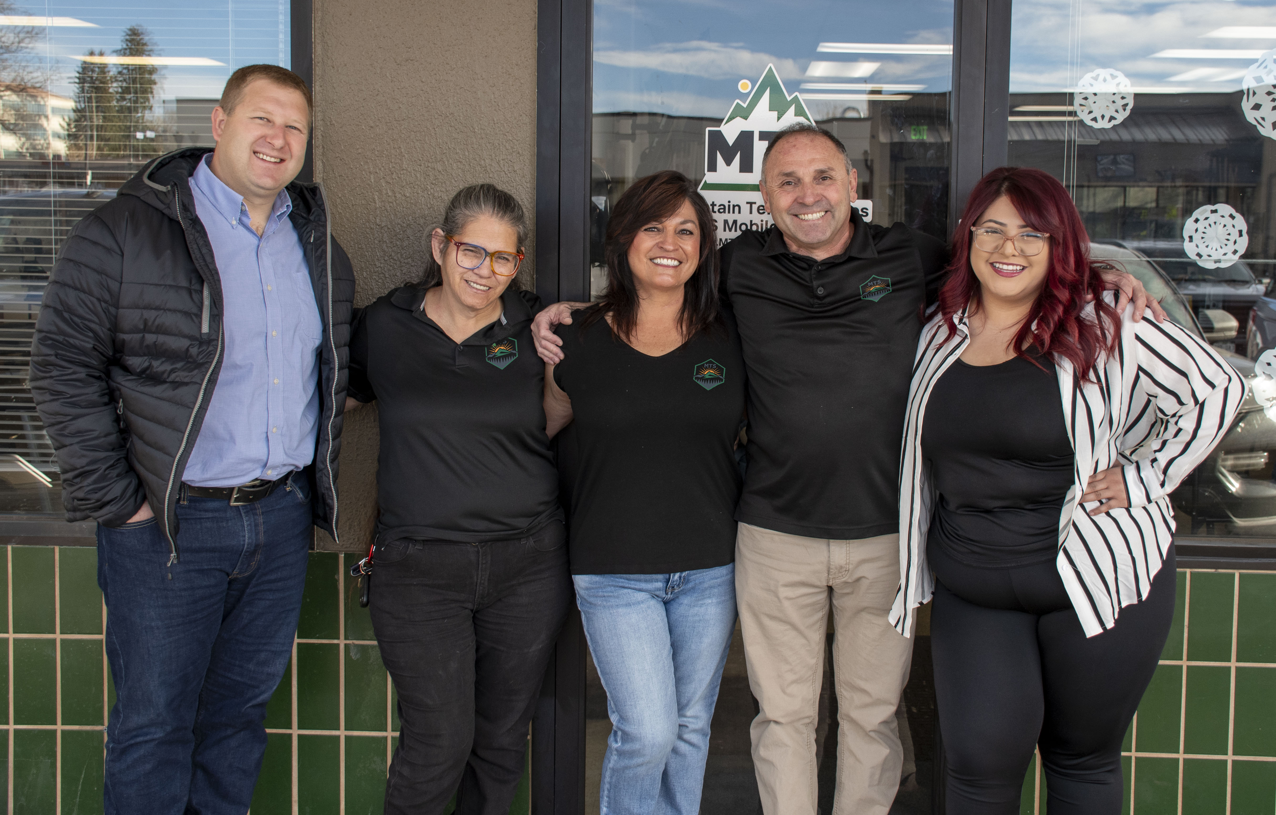 Group of five people standing in front of a glass door, smiling, with some wearing black shirts with a mountain logo.