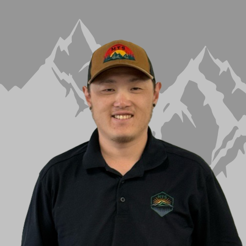 A young man wearing a tan cap with a mountain logo and a black collared shirt with a mountain logo on it, smiling at the camera, standing in front of a gray background with a white mountain silhouette.