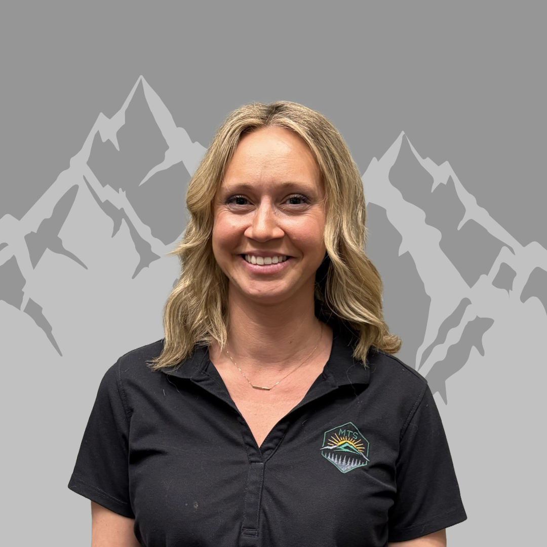 A smiling woman with blonde hair wearing a black shirt with a Mountain technical services logo, standing in front of a gray background with mountain outline graphics.