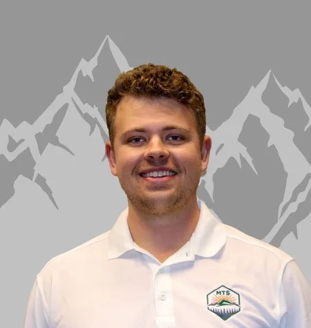Young man with curly hair and a beard smiling, wearing a white polo shirt with a logo, standing in front of a mountain-themed background.