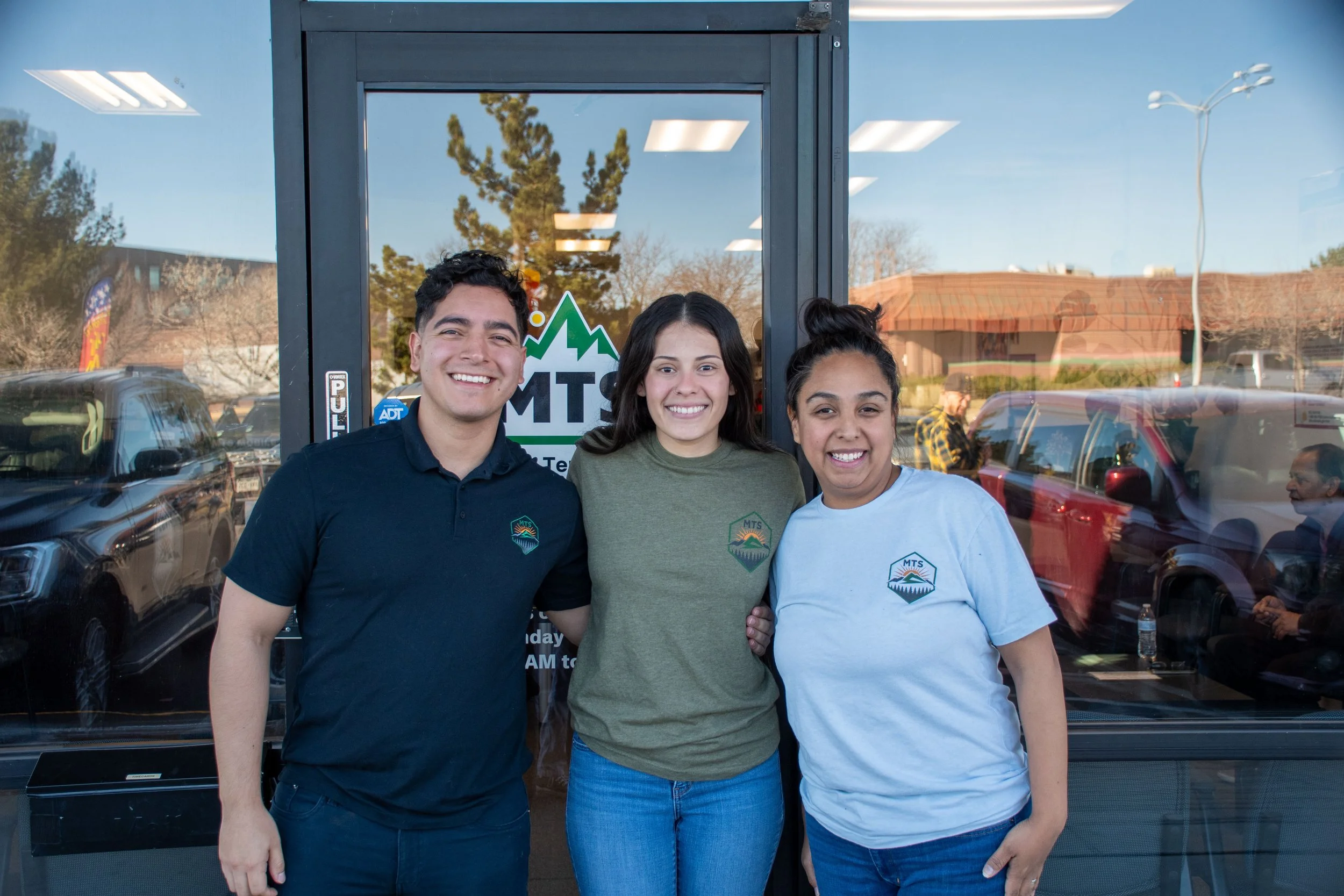 Three smiling young adults standing outside a building with glass door, wearing casual clothing with a logo, reflecting a parking lot and trees in the background.
