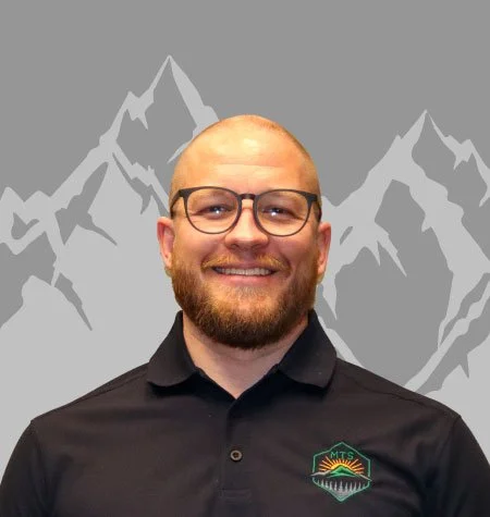 A smiling man with glasses and a beard wearing a black polo shirt with a mountain-themed logo, standing in front of a gray mountain backdrop.