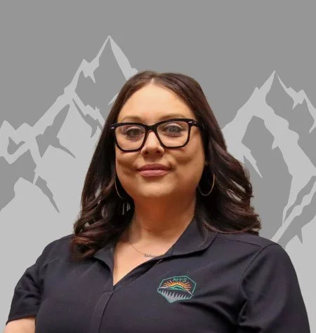 A woman with dark hair, glasses, and hoop earrings, wearing a black polo shirt with a mountain logo, stands in front of a mountain image background.