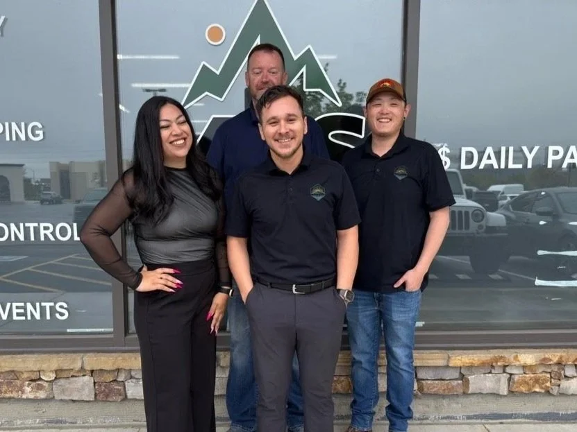 Four people, three men and one woman, standing outdoors in front of a glass building with a mountain and financial graph design behind them, smiling for the photo.