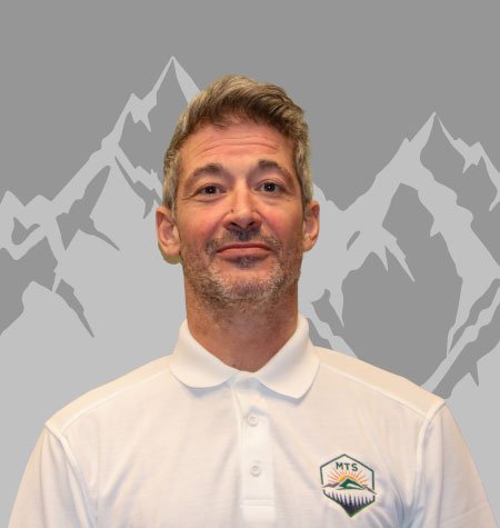 Man with short light brown hair and a beard, wearing a white polo shirt with an MTS logo, standing in front of a mountain-themed background.