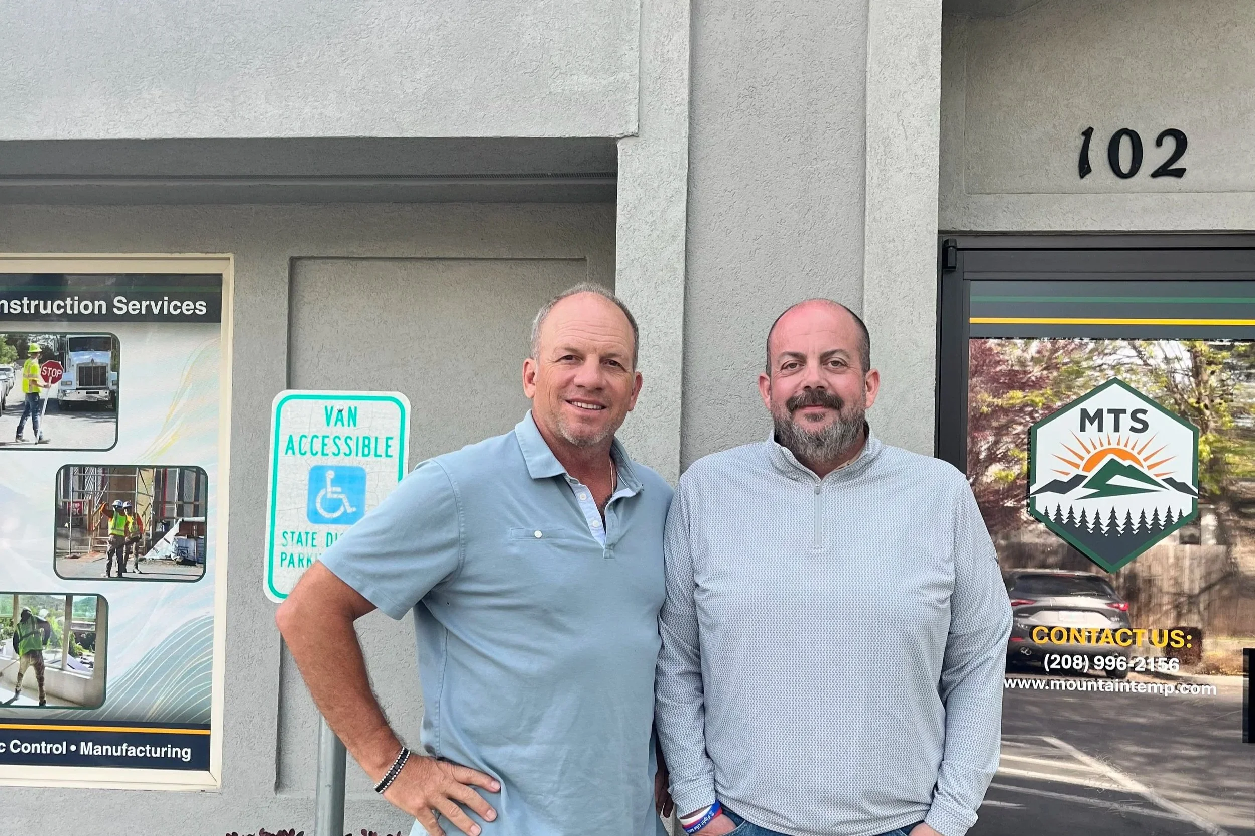Two men standing outside a building with construction or business signage, smiling at the camera.