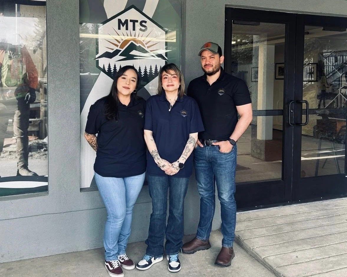 Three people standing outside a building with a logo that has mountain and forest imagery, wearing black polo shirts with a small logo on the left chest.
