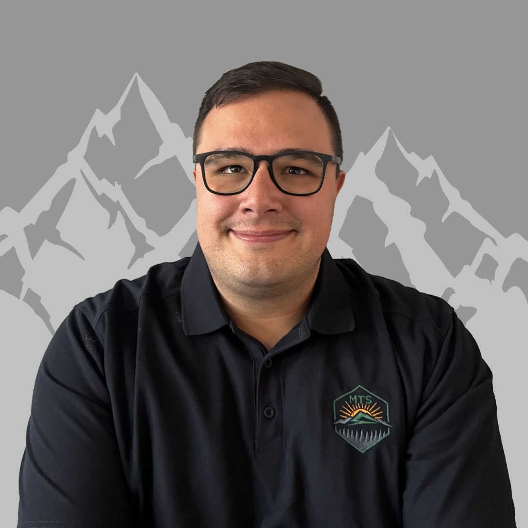 A man with glasses and short dark hair wearing a black polo shirt with a mountain and sunrise logo, standing in front of a digital background of gray mountains.