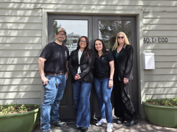 Four people standing in front of a building with a glass door, smiling. Two women and two men, casually dressed, with the women wearing black blazers and jeans, and the man wearing a black t-shirt and jeans, outside on a sunny day.