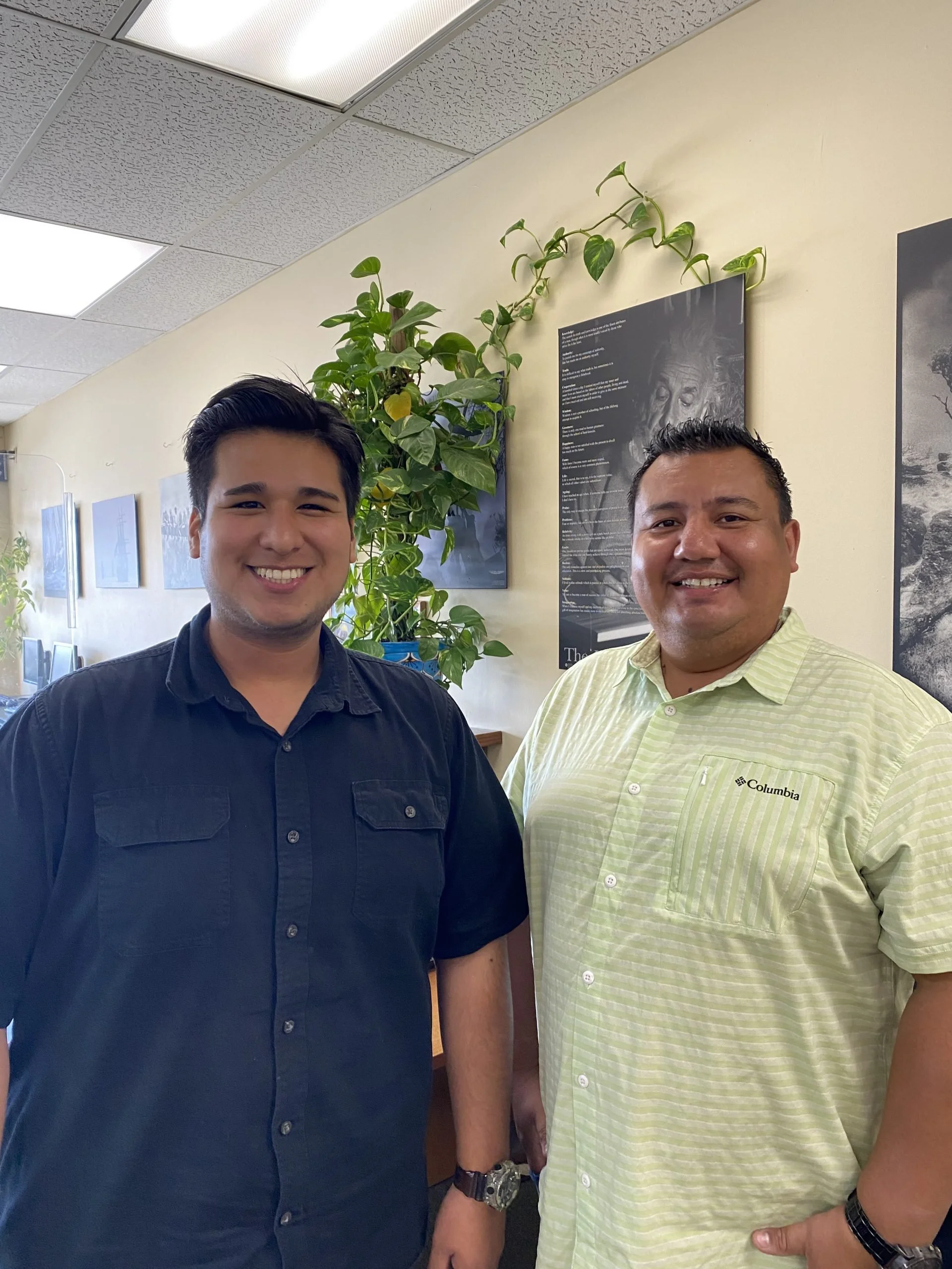 Two men standing indoors, smiling at the camera with a green leafy plant behind them and framed artwork on the wall.
