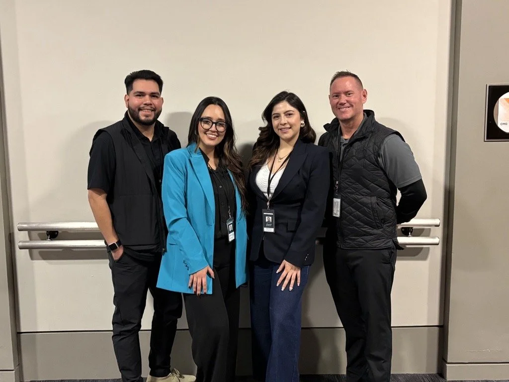 Four people standing together indoors in front of a wall. They are smiling and wearing conference badges.