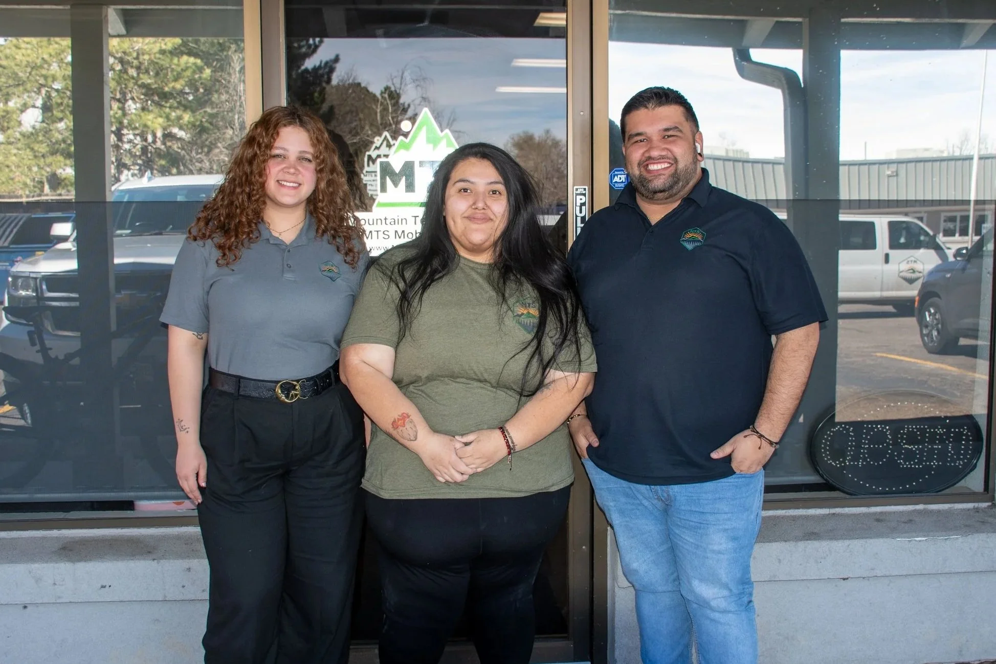 Three people standing side by side in front of a glass door, smiling at the camera, outside a building labeled Mountain TMS Mobile.