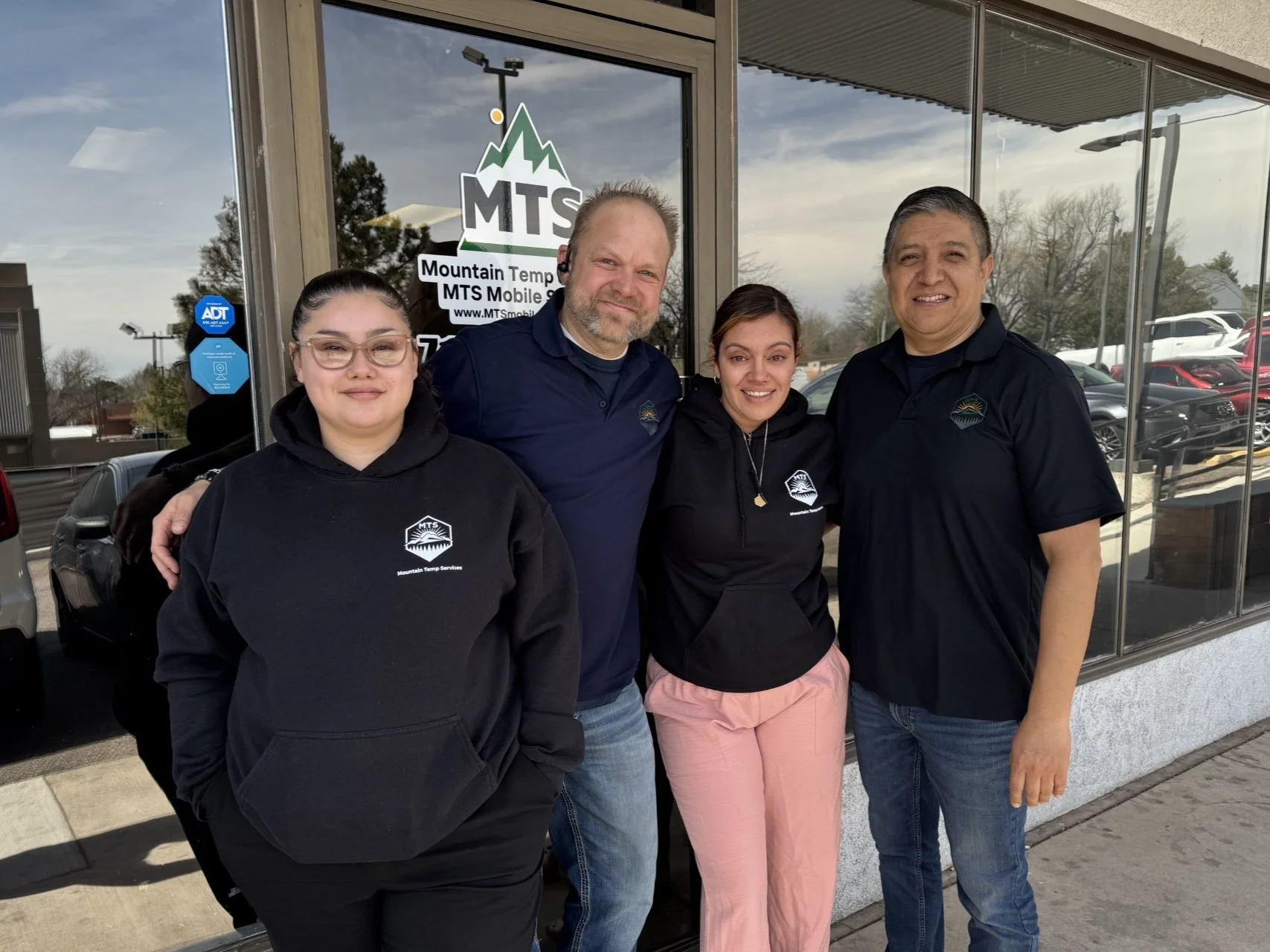 Four people standing outside a building with large glass windows, smiling for the camera. The door has a sign for Mountain Temp Services (MTS), indicating the location. The group includes two women and two men, all wearing dark-colored clothing, some with logos related to Mountain Temp Services.
