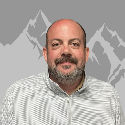 A man with a beard and mustache, wearing a zip-up patterned jacket, standing in front of a gray background with mountain illustrations.