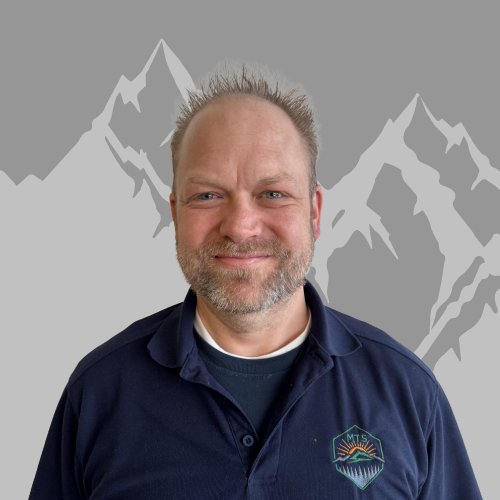 A man with short, spiky hair and a beard smiling, wearing a navy blue collared shirt with a logo on the chest, standing in front of a digitally illustrated mountain background.