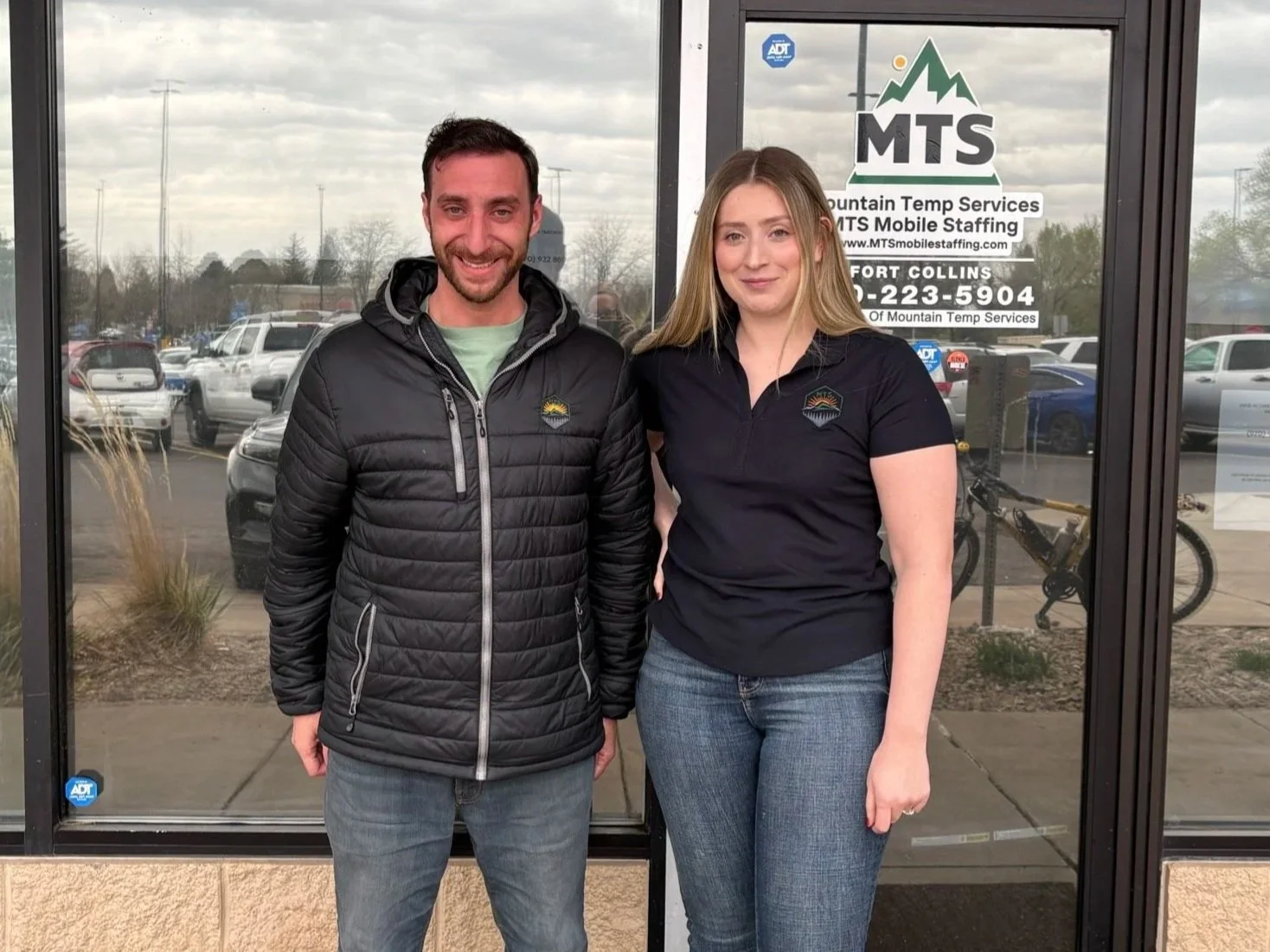 Two people standing outside a glass door, smiling, with a parking lot in the background. Both are wearing black jackets with a mountain logo on the chest.