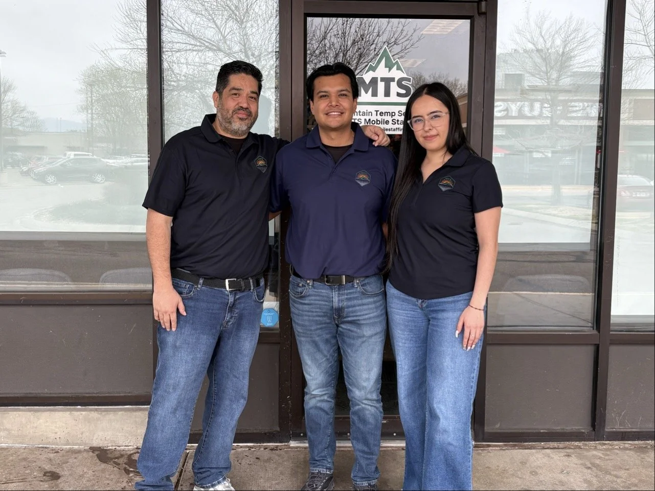 Three people standing side by side in front of a glass door with a sign that reads 'MTS Mountain Temp Mobile' and trees visible in the background. They are dressed in matching dark blue polo shirts with a logo.