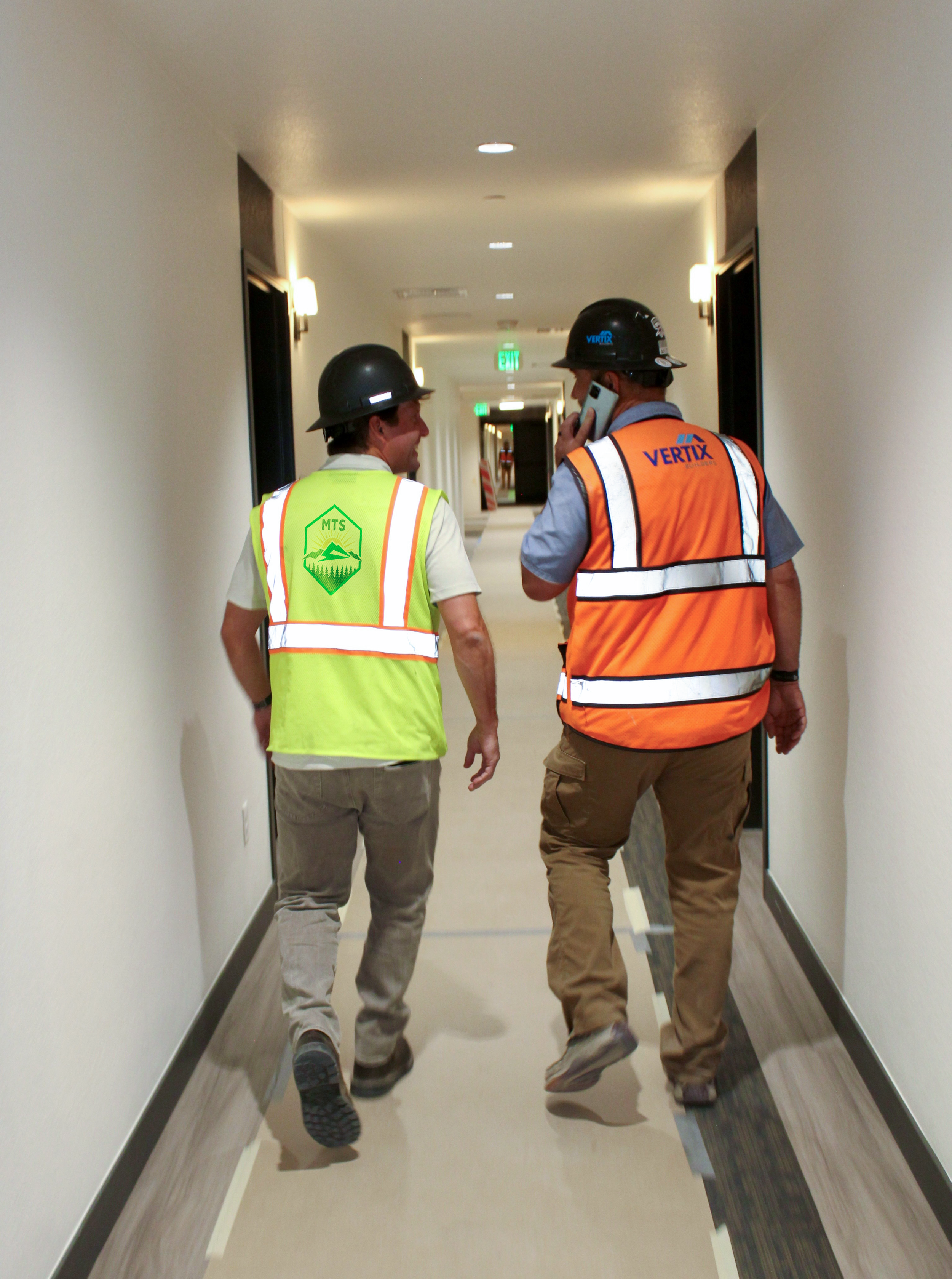 Two construction workers walking down a hallway while talking on a cell phone. They are wearing safety helmets and vests, one in green and the other in orange, with construction branding.
