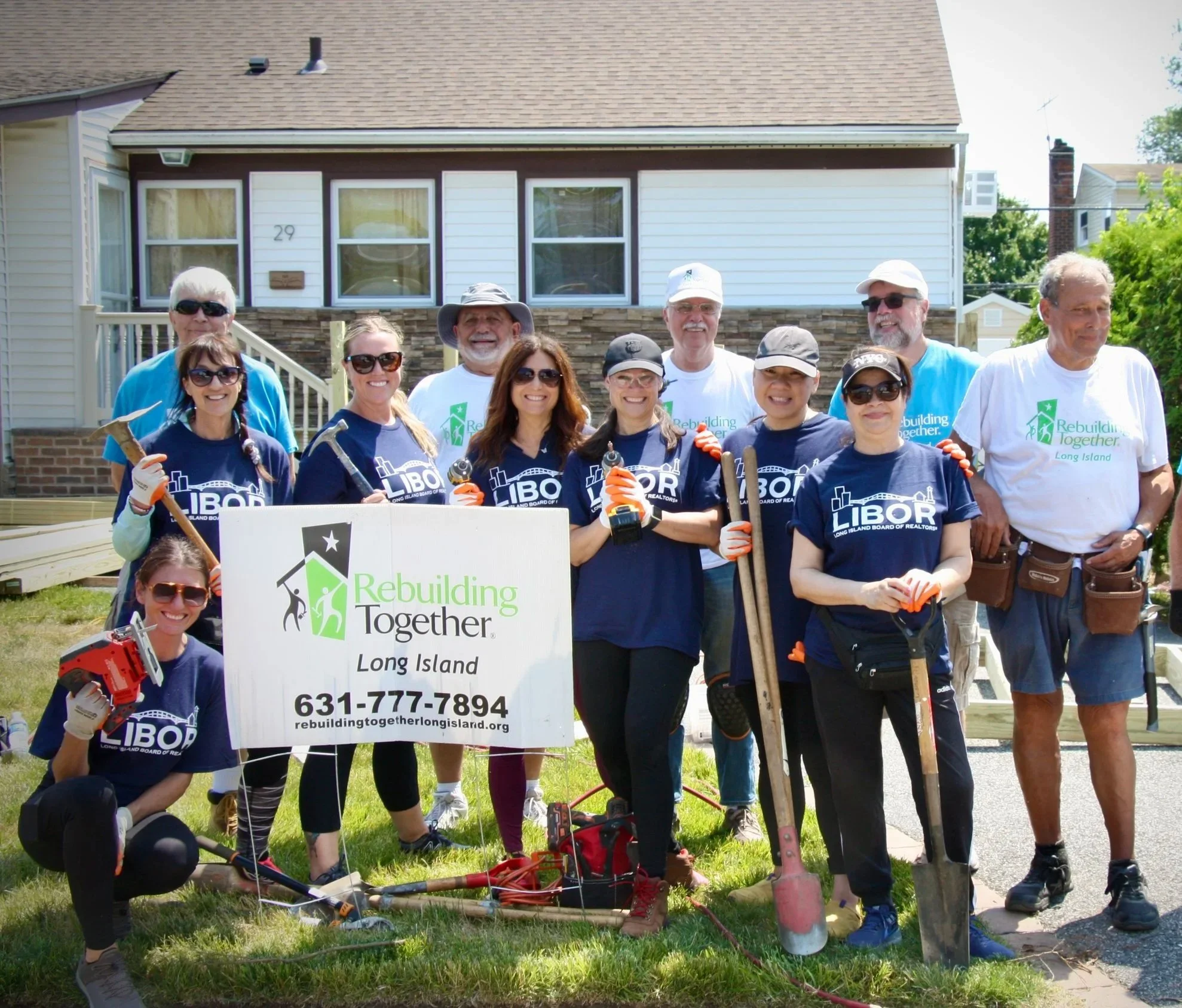 Twelve smiling male and female volunteers holding Rebuilding Together Long Island sign in front yard of job site