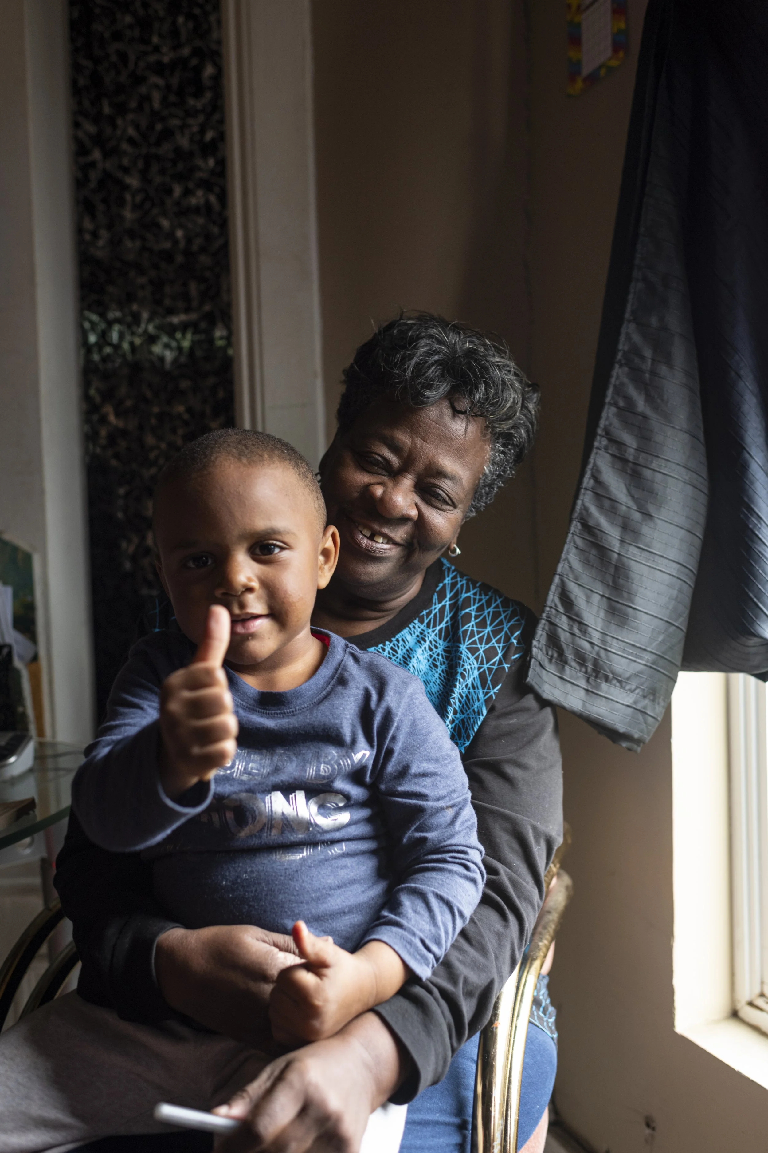 Child in blue shirt smiling while sitting on smiling grandmother's lap while he gives a thumbs up