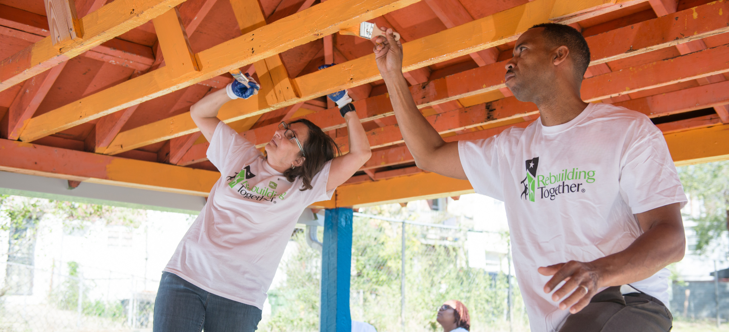 A woman and a man wearing white tee shirts with Rebuilding Together logo paint the rafters of a covered porch with yellow paint.