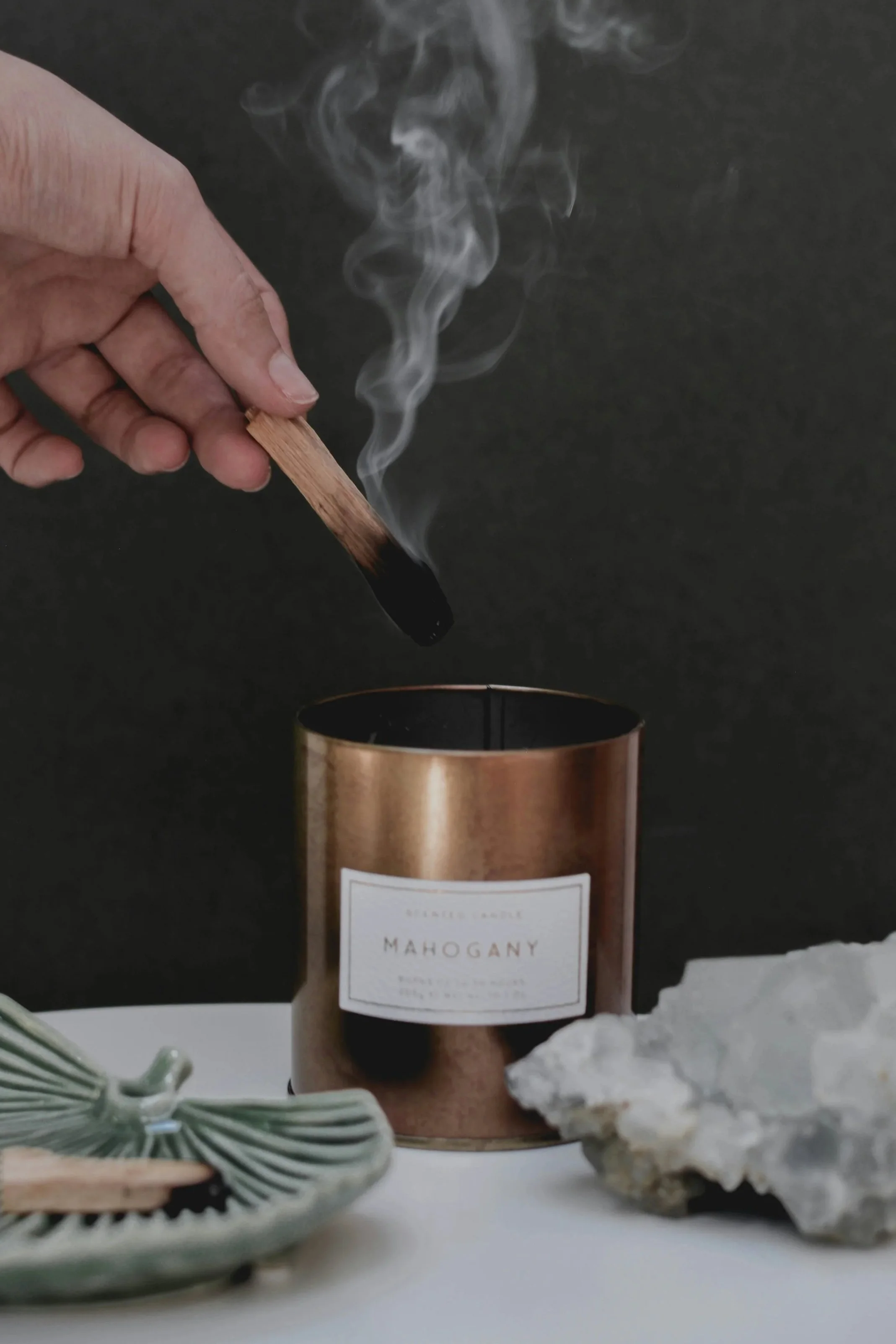 Hand holding a lit wooden palo santo stick above a candle labeled 'Mahogany' on a white surface with a decorative ceramic dish and a rock nearby. Palo santo recenter energy for acupressure healing while in Easton, MD