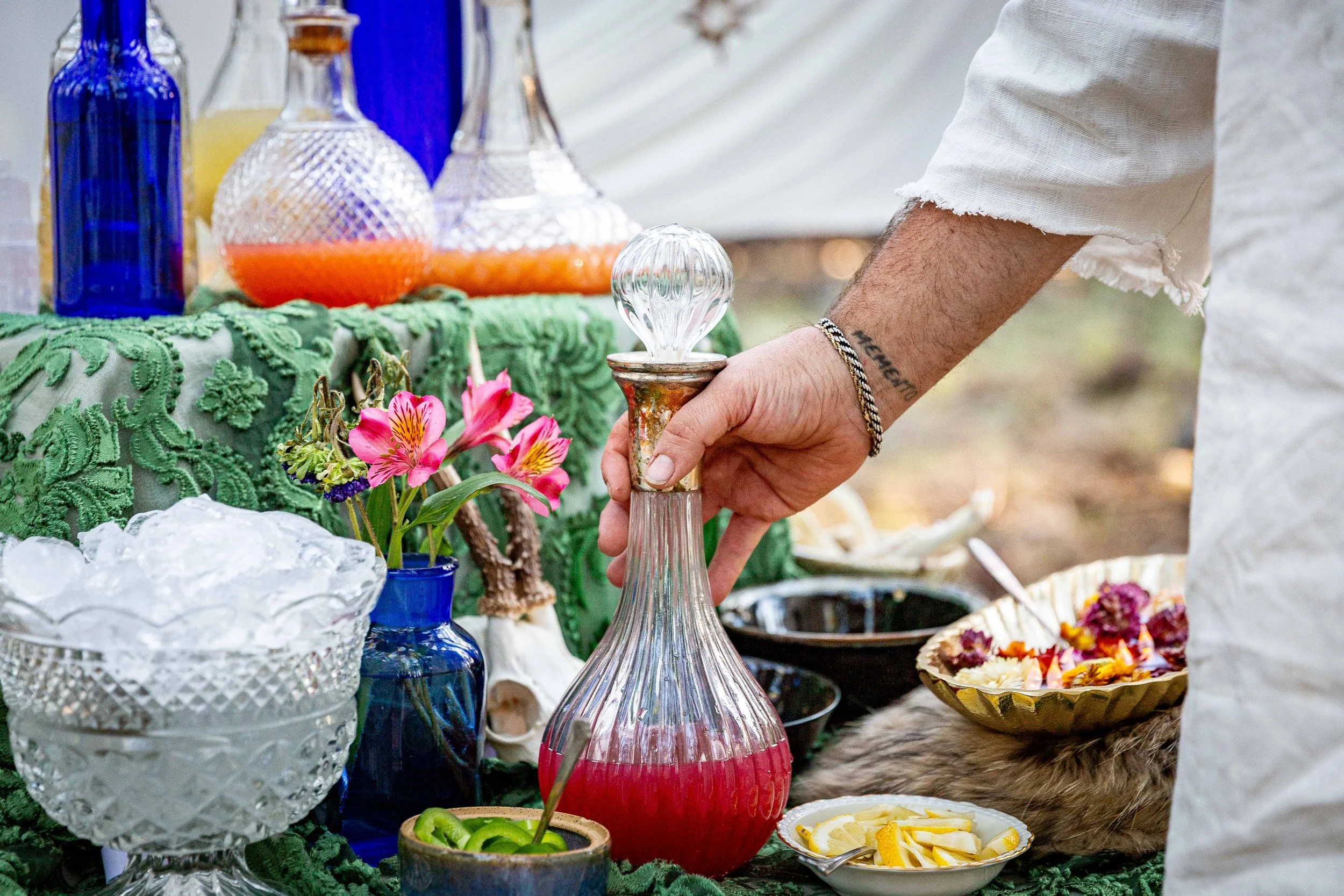 Person holding a glass decanter with a wooden stopper at an outdoor table decorated with colorful glass bottles, flowers, and bowls of food.
