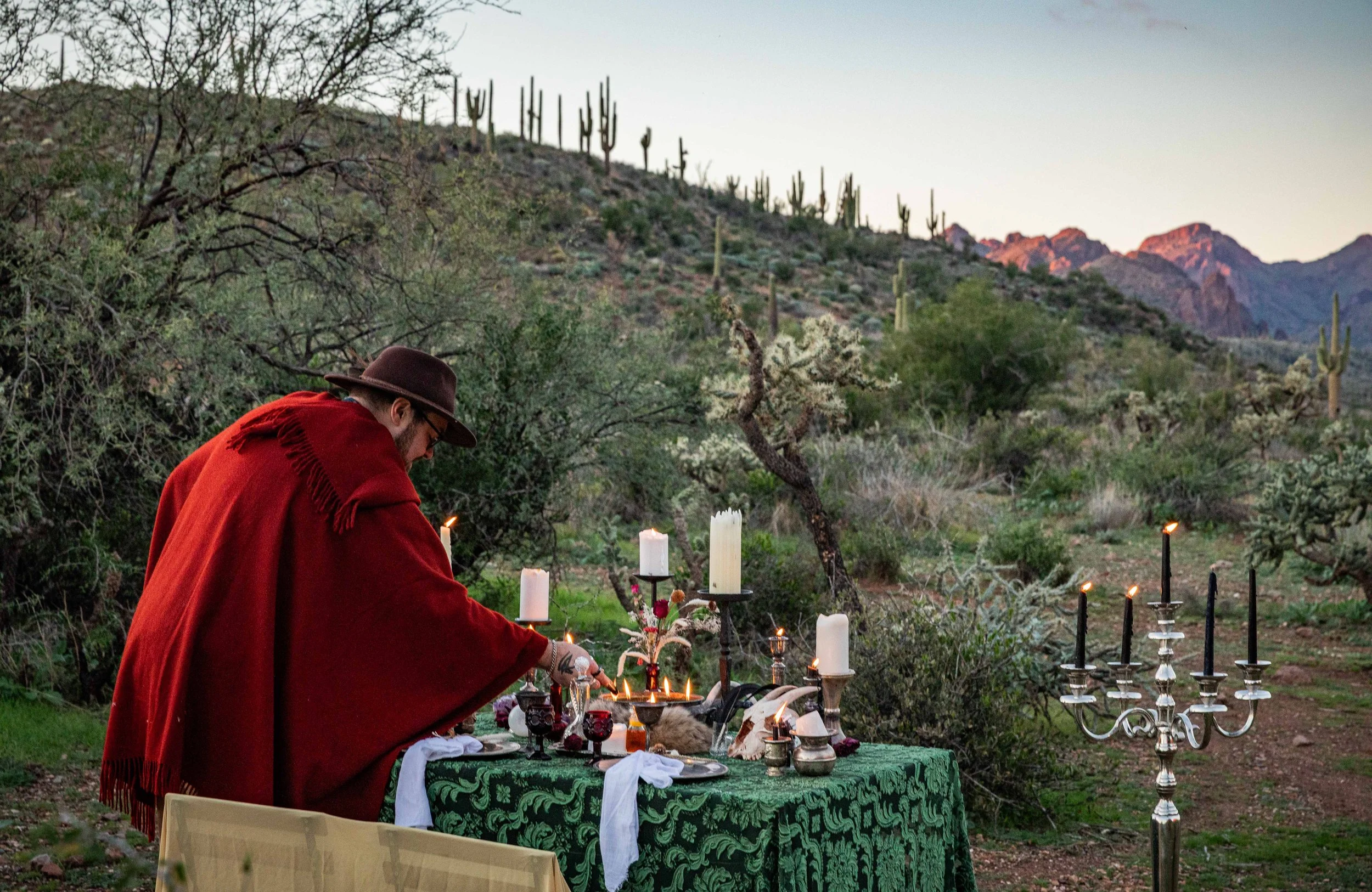 A man in a red cape and a brown hat setting candles on a decorated outdoor table in a desert landscape with cacti and mountains in the background.