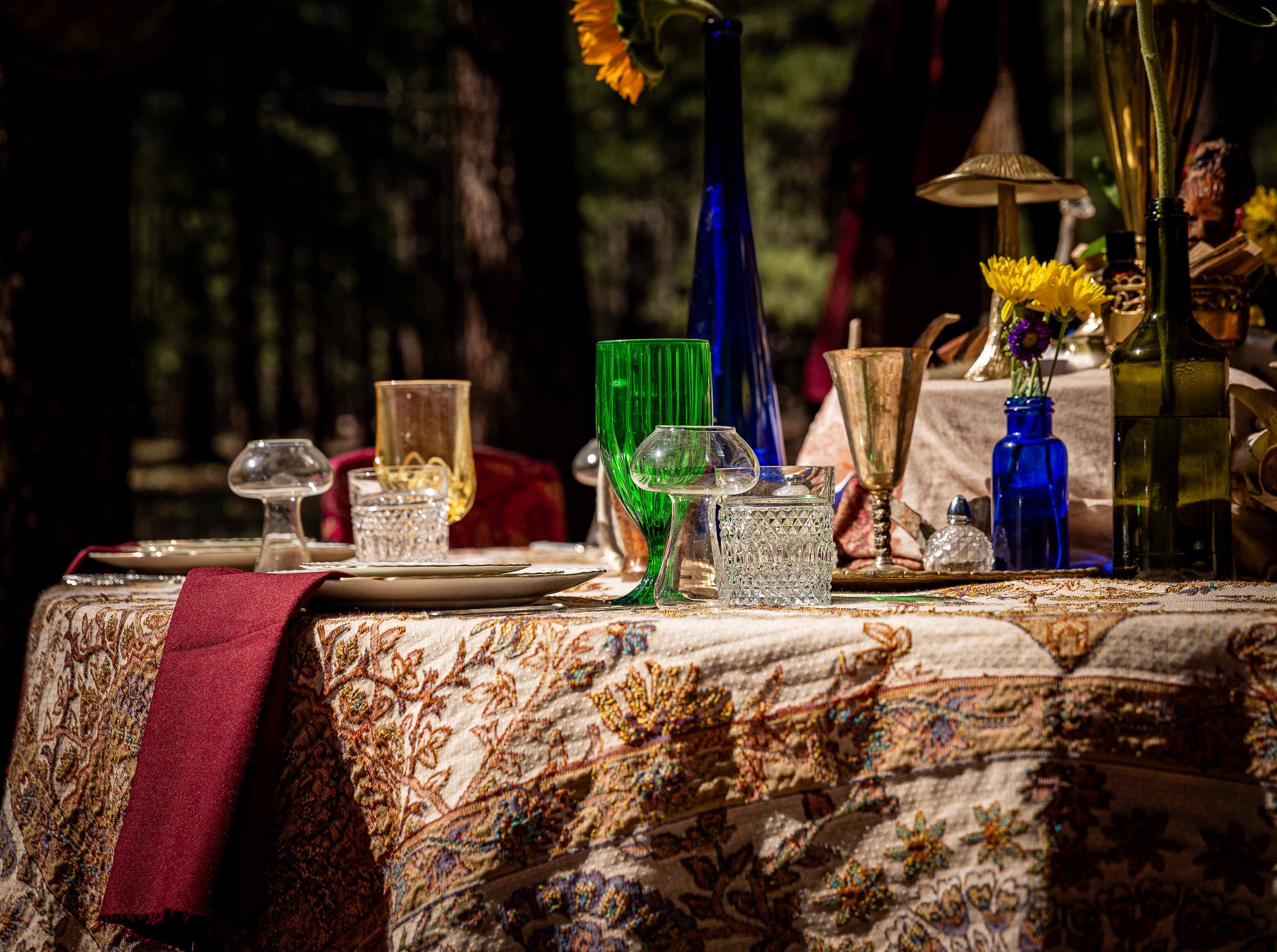 A table with a floral tablecloth set outdoors with colorful glass vases and dishes, with a red cloth draped over one side, surrounded by trees in the background.