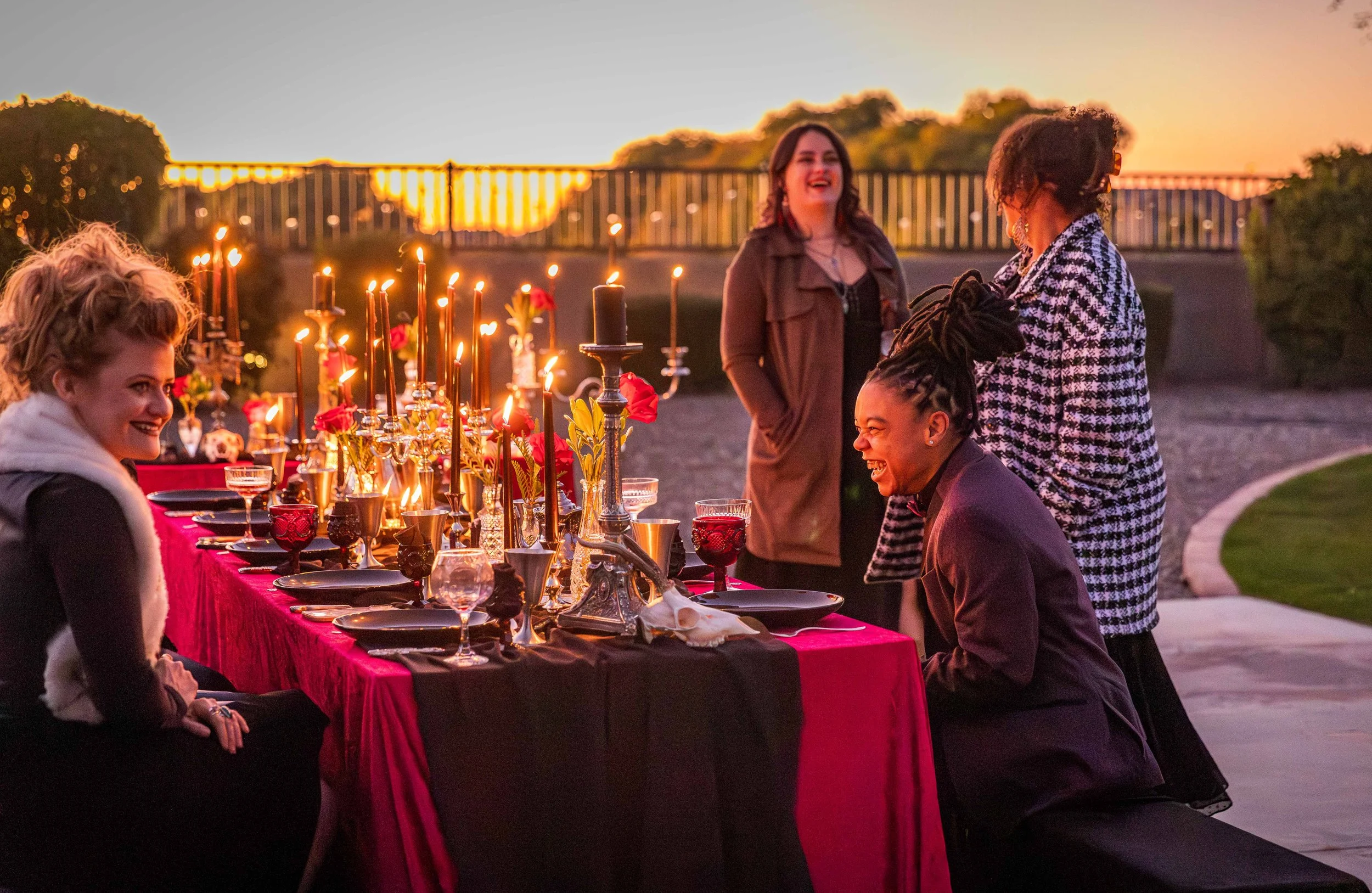 People celebrating a dinner party outdoors during sunset with a table set with candles, flowers, and glassware, engaging in conversation and laughter.