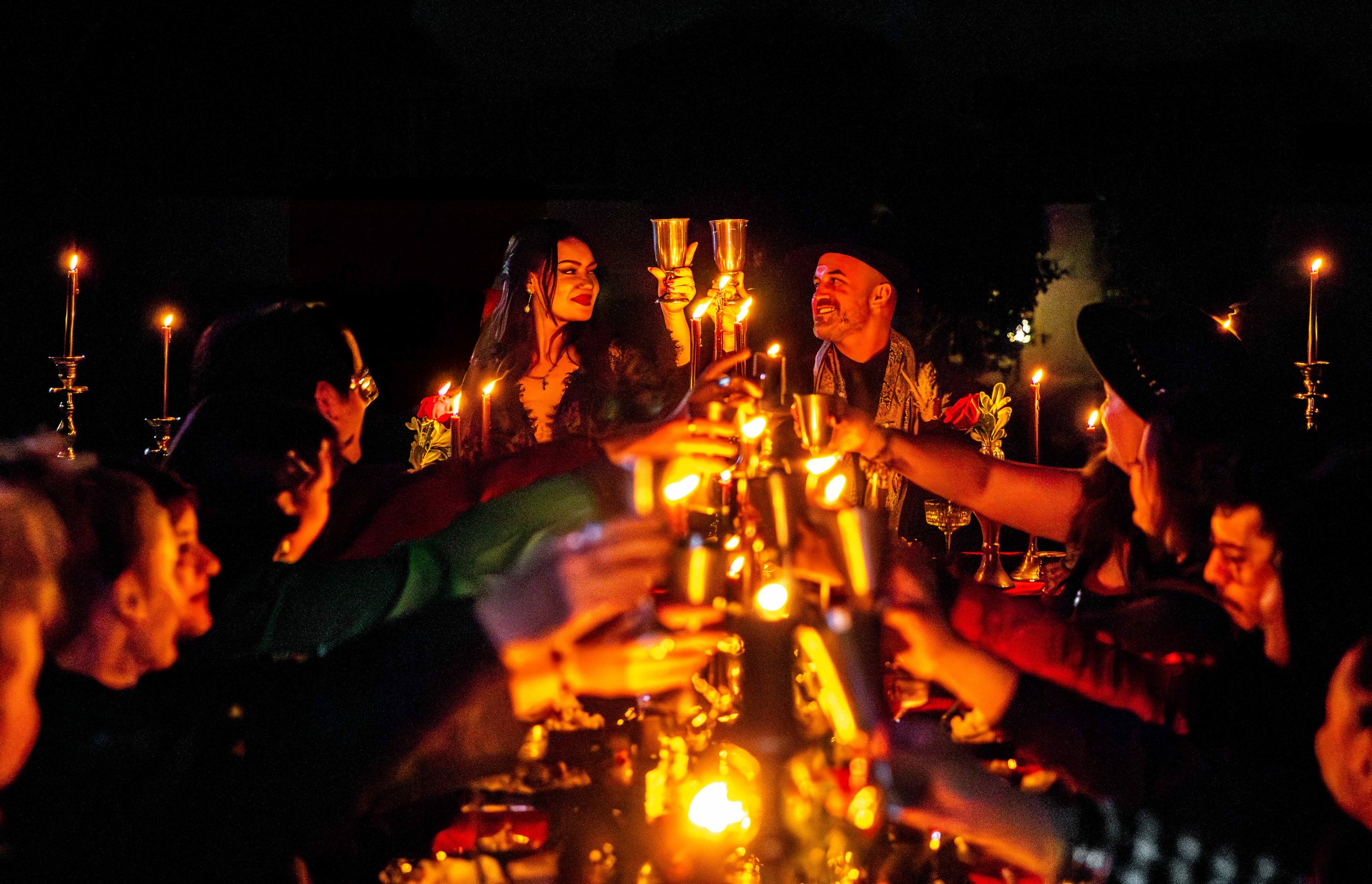People gather around a long table outdoors at night, raising glasses in a toast, with lit candles and candle holders creating a warm festive atmosphere.