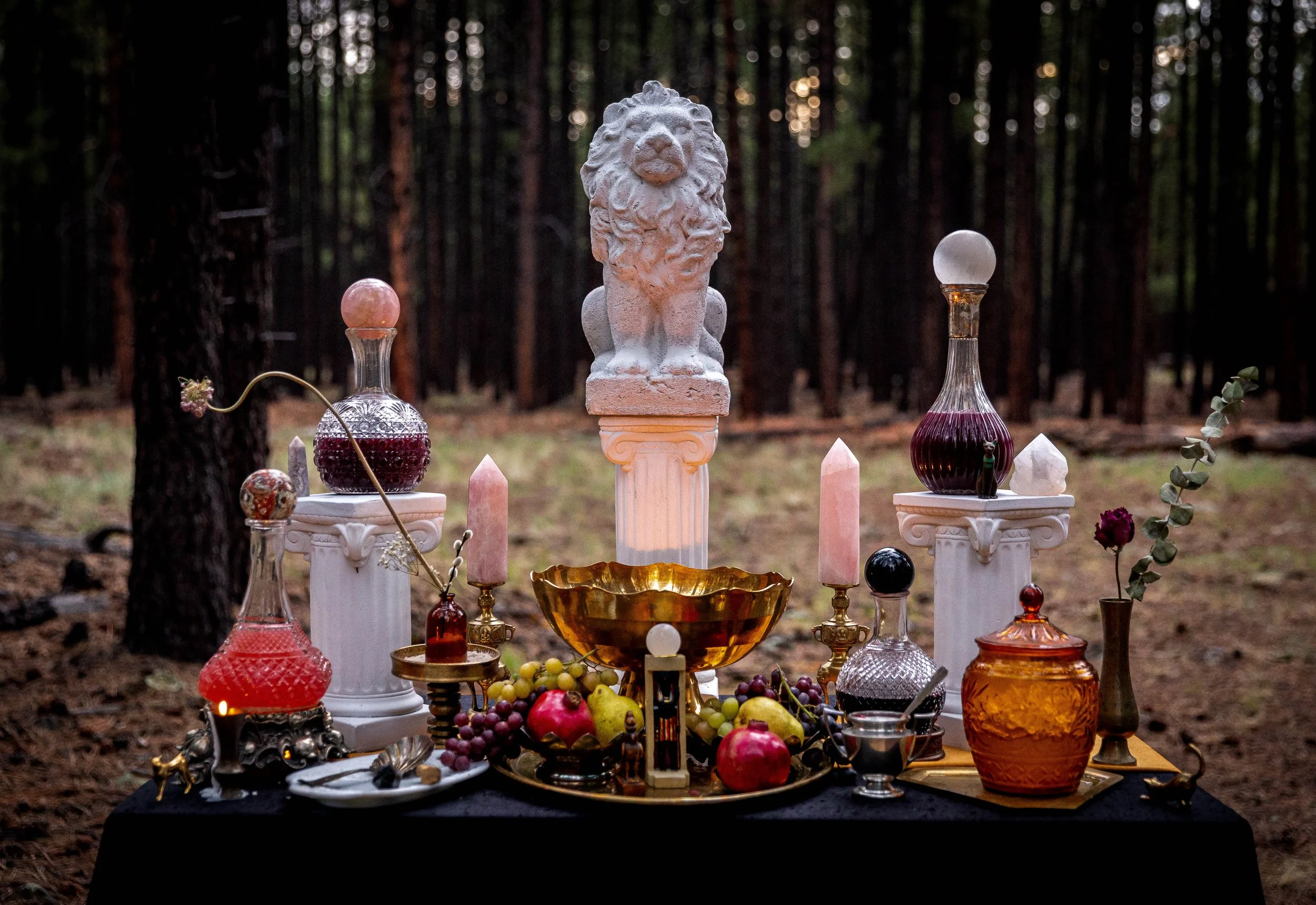 An altar in a forest with various glass bottles, candles, fruits, and decorative items, including a white lion sculpture on a pedestal.