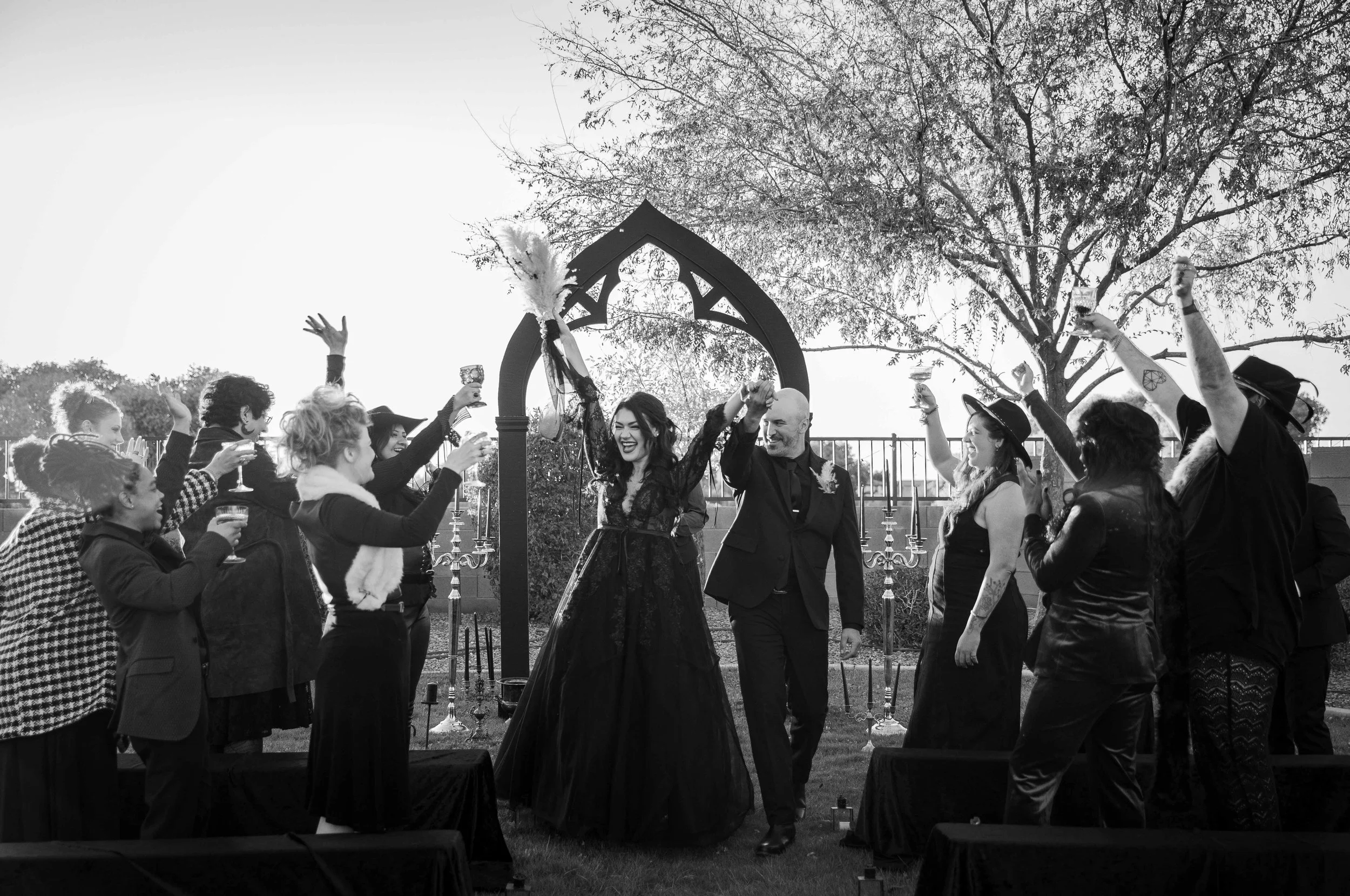 A wedding celebration outdoors with the bride and groom holding hands and smiling, surrounded by friends raising glasses in toast, under a decorative arch and trees.
