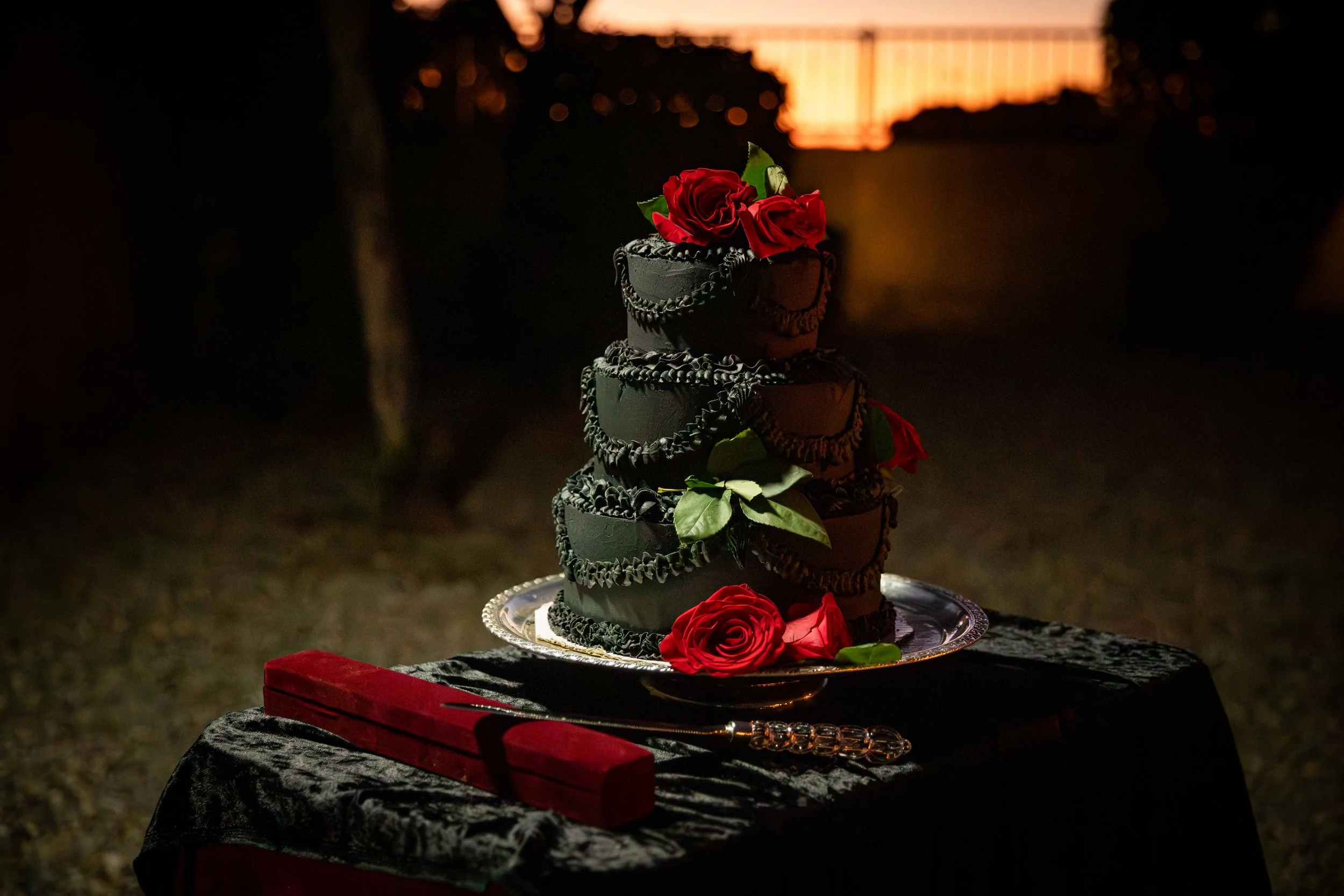A four-tier black wedding cake decorated with red roses and green leaves, placed on a silver tray on a black tablecloth. A red brick-shaped object and a decorative knife are also on the table. The background is dark with a sunset or fire glow visible in the distance.
