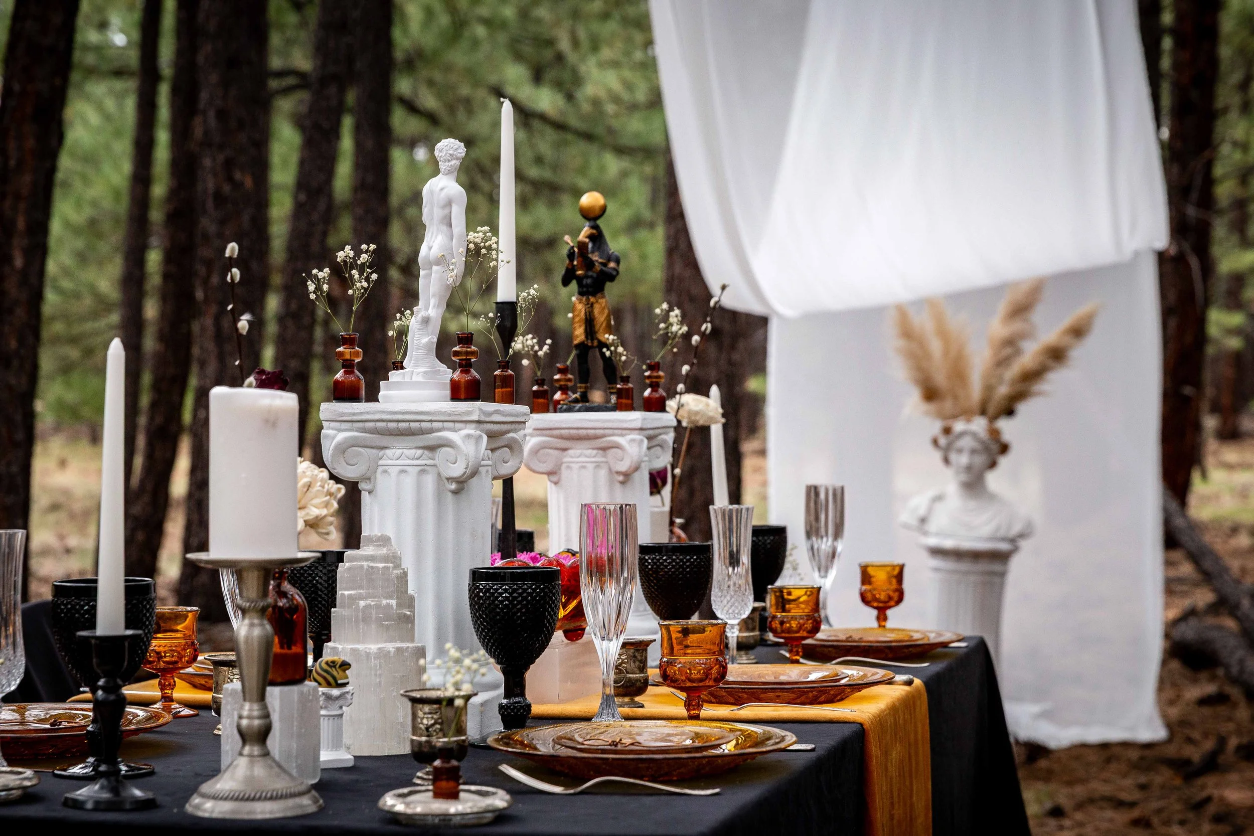 Table set for an outdoor event with decorative statues, candles, and tableware in a forest setting.