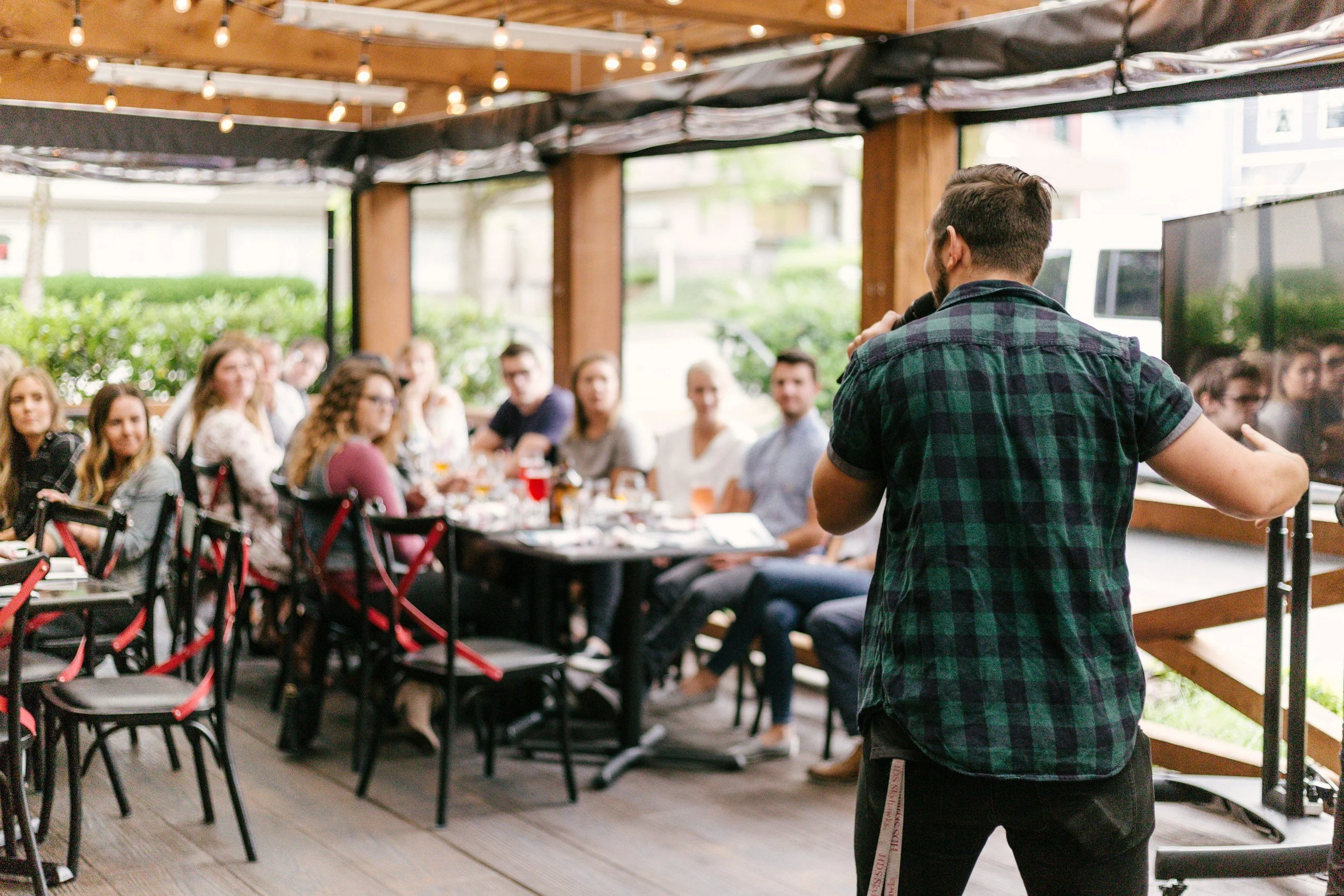 A man in a plaid shirt gives a speech into a microphone in front of a seated audience at a restaurant or cafe with string lights overhead.
