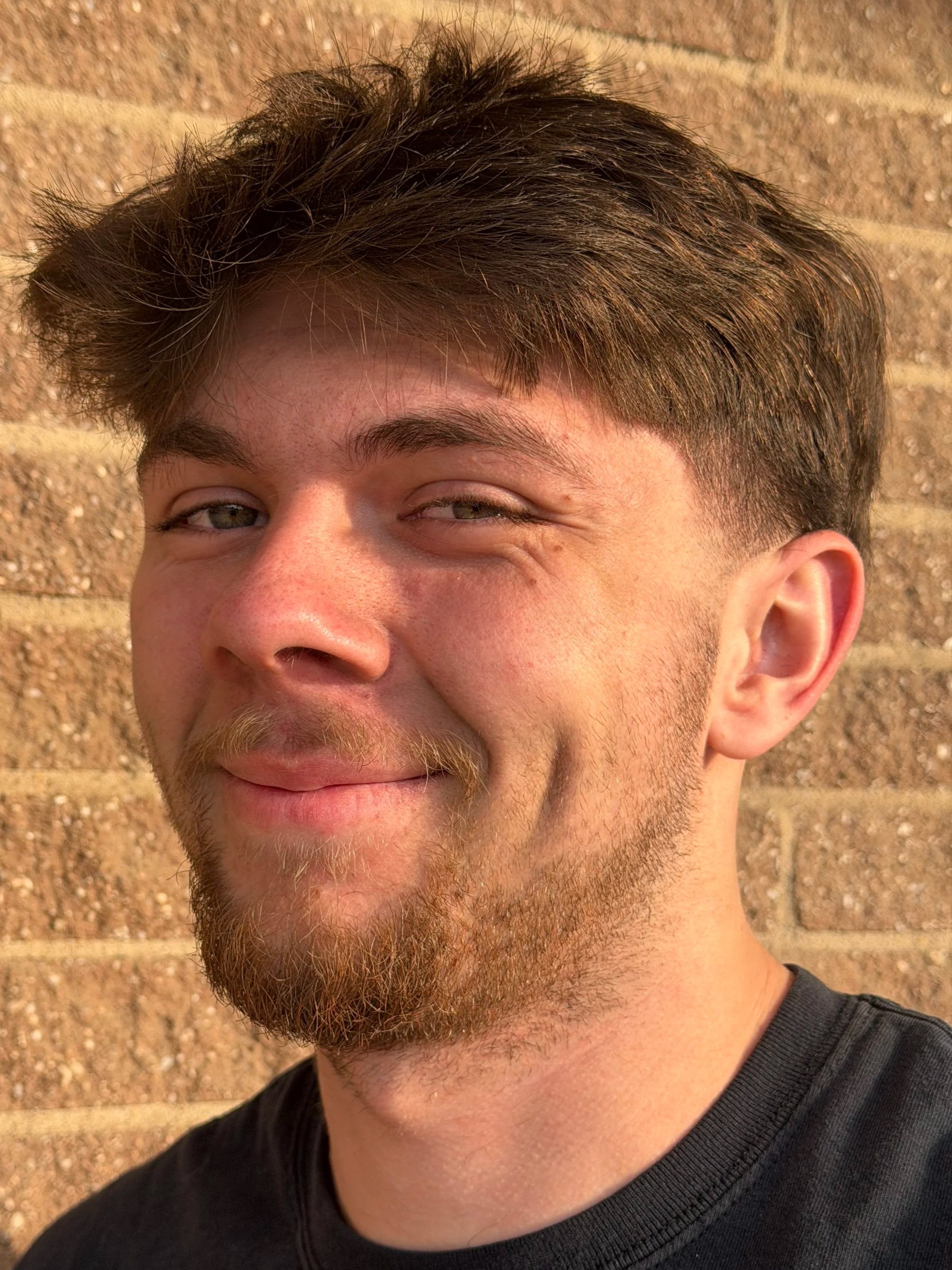 A young man with brown hair and a beard, smiling with a wink, standing against a brick wall.