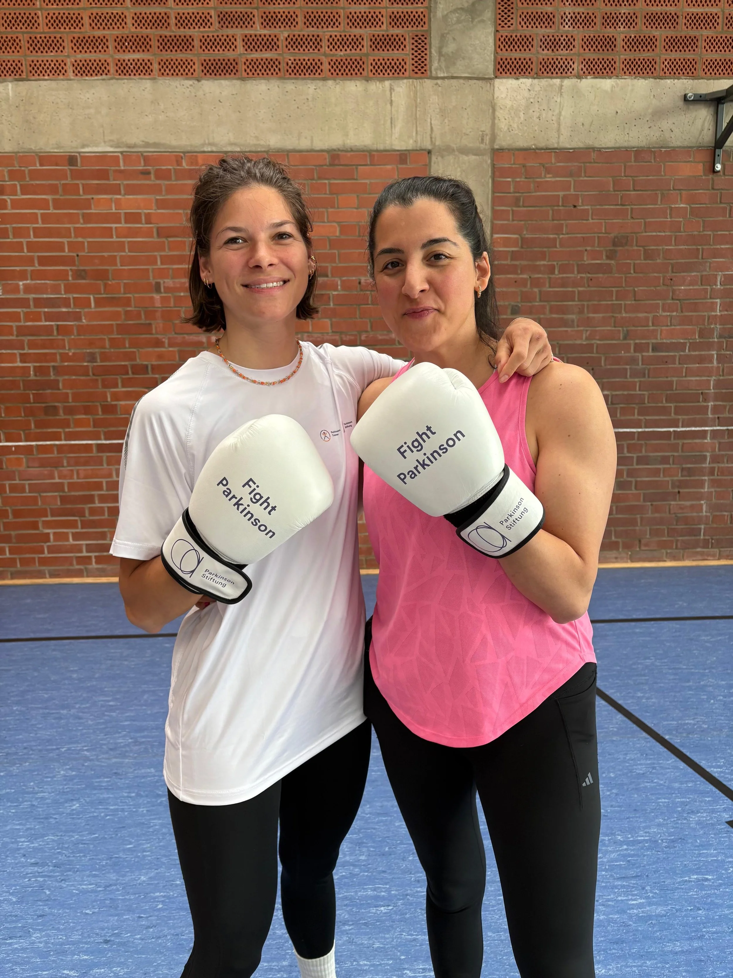 Zwei Frauen in Sportkleidung tragen Boxhandschuhe mit der Aufschrift "Fight Parkinson" und posieren im Fitnessraum.