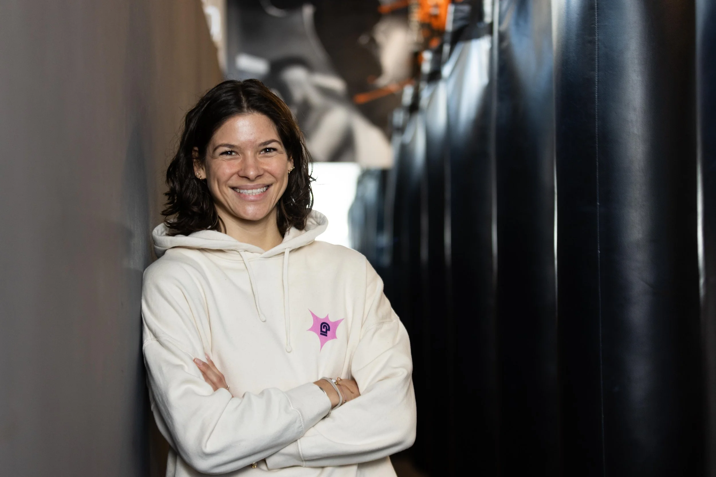 Junge Frau mit braunen, lockigen Haaren trägt weißen Hoodie mit rosa Logo, steht in einem Raum zwischen schwarzen Boxsäulen, lächelt, Arme verschränkt.