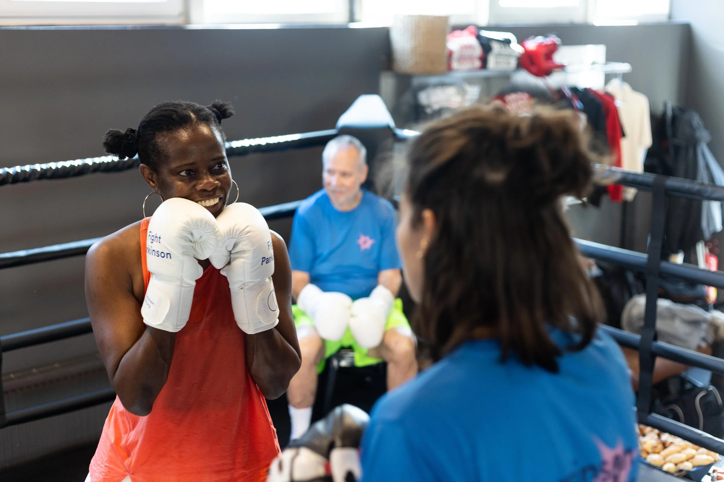Eine junge schwarze Frau in roten Boxhandschuhen und Trainingskleidung hält in einem Boxring bereit, während eine andere Frau mit braunen Haaren, in blauer Kleidung, mit ihr spricht. Im Hintergrund sitzt ein älterer Mann in sportlicher Kleidung auf e