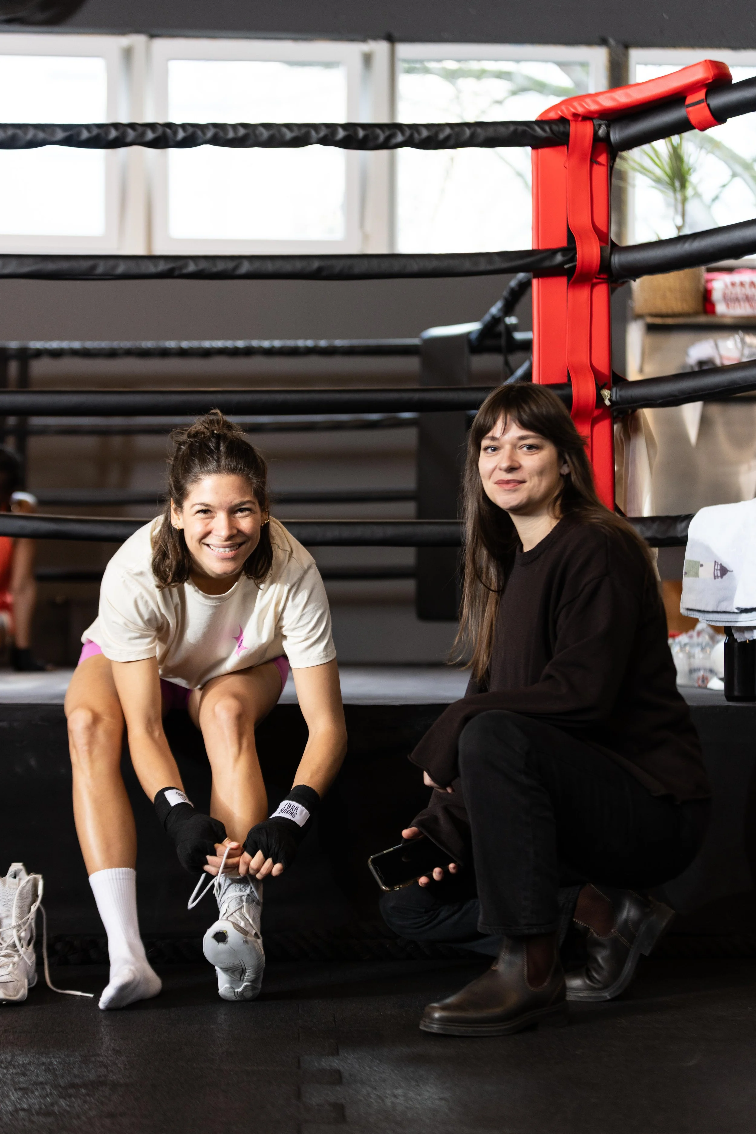 Zwei Frauen sitzen vor einem Boxring in einem Fitnessstudio, eine lächelnd beim Anziehen ihrer Turnschuhe, die andere sitzt neben ihr und hält ein Smartphone.