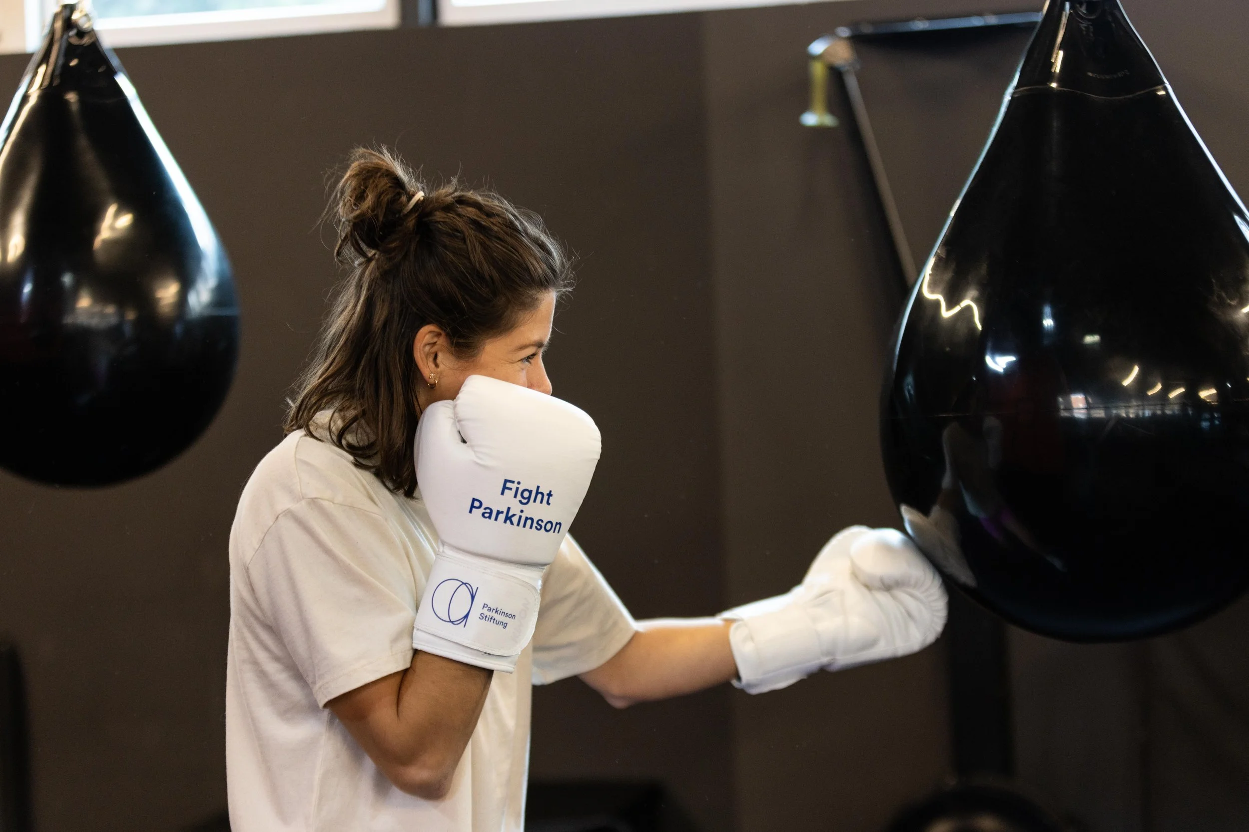 Eine Frau trägt Boxhandschuhe mit der Aufschrift 'Fight Parkinson' beim Training an einem schwarzen Boxsack.