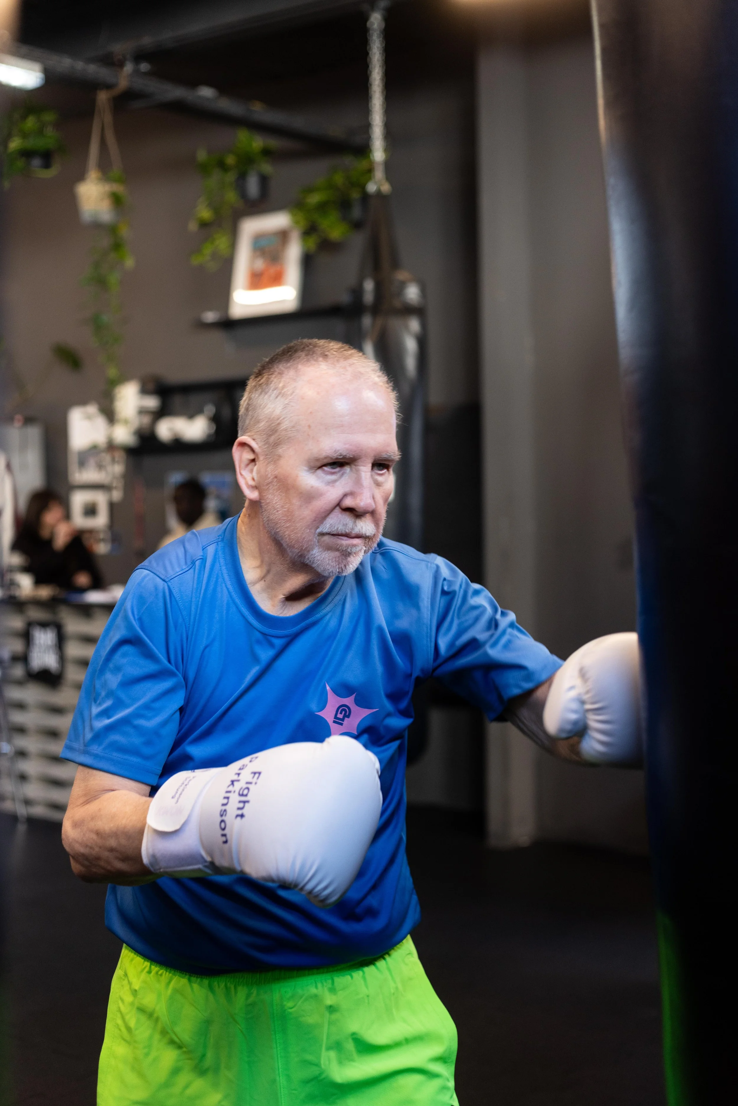 Älterer Mann mit Parkinson trainiert im Fitnessstudio in Frankfurt am Main mit Boxhandschuhen, beugt sich vor und boxt gegen einen schwarzen Boxsack.