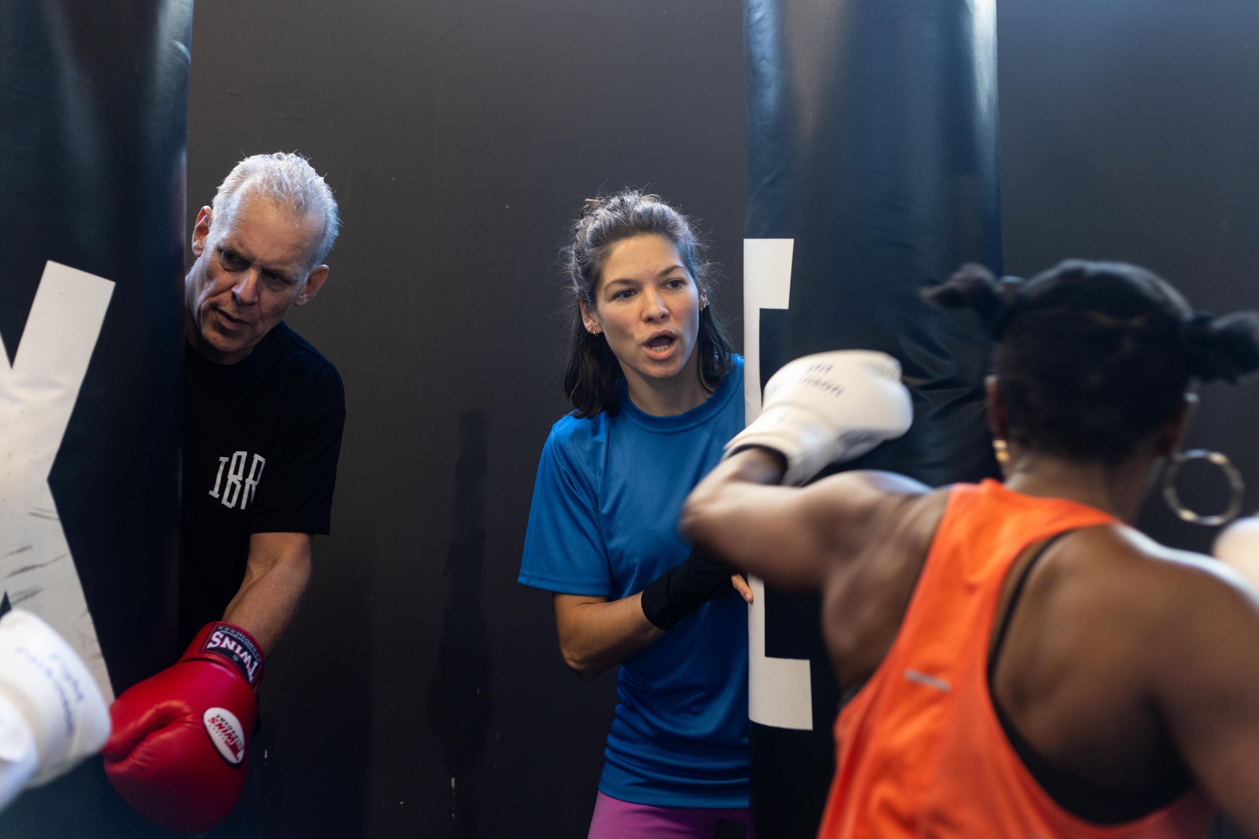Trainer erläutert Technik bei Boxtraining, eine Frau im blauen T-Shirt, eine andere in einem orangefarbenen Top und Boxhandschuhen, eine weitere Person mit grauem Haarschopf beobachtet. Die Gruppe ist an Parkinson erkrankt und ist in Frankfurt am Mai