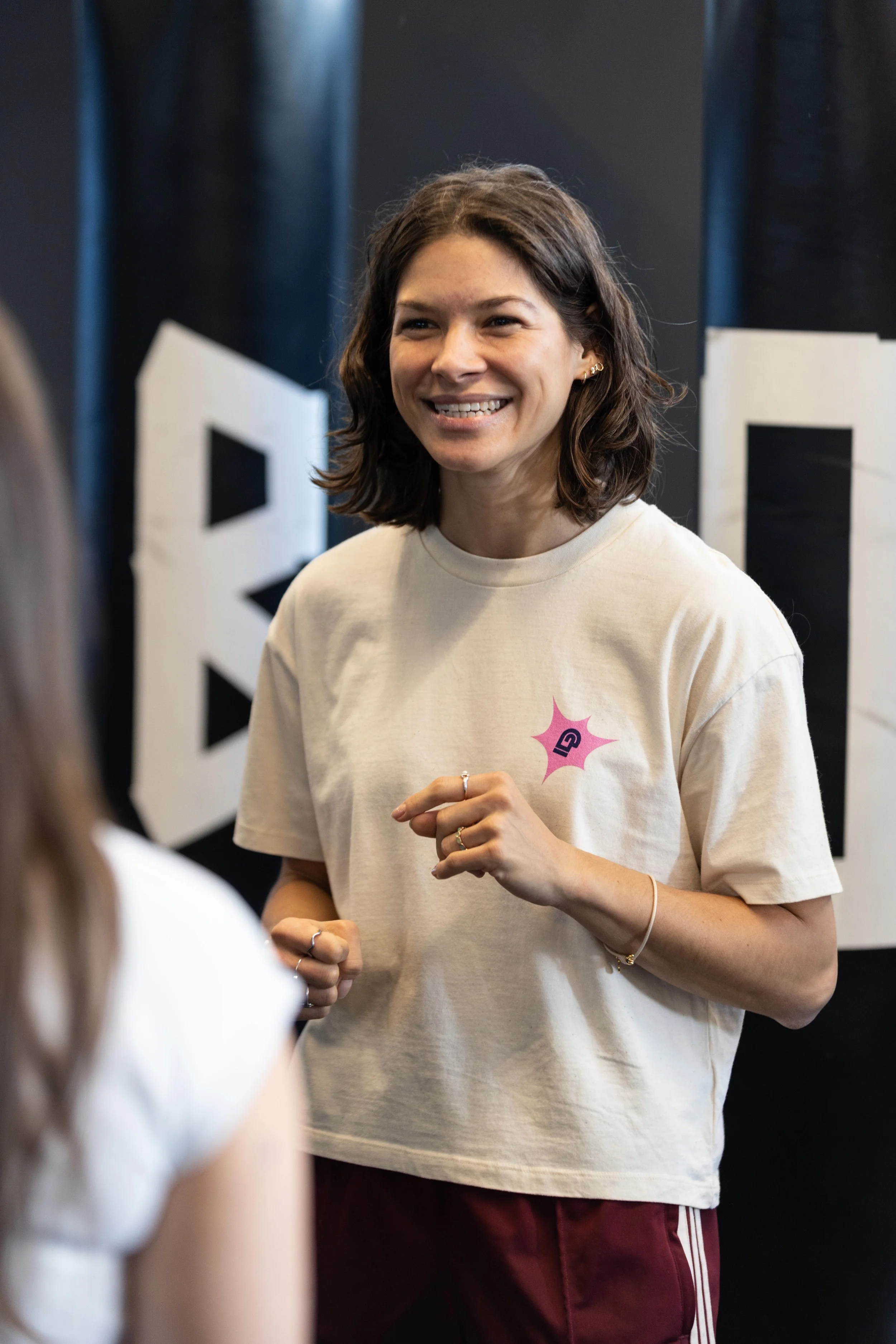 Eine lächelnde Frau mit braunen, schulterlangen Haaren trägt ein weißes T-Shirt mit einem pinken und blauen Logo und steht in einem Raum mit dunklem Hintergrund, in dem große Buchstaben zu sehen sind.