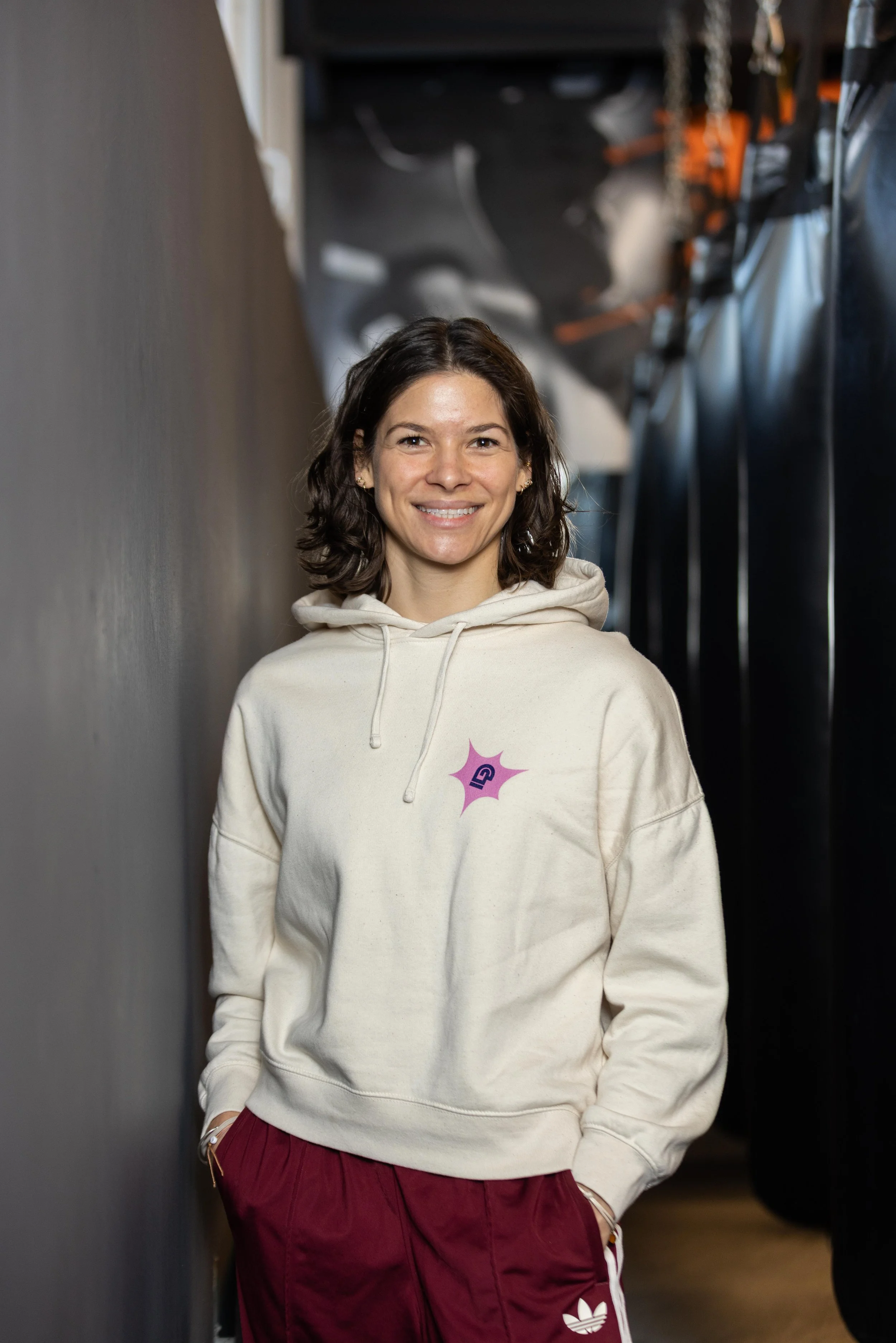 Eine lachende Frau in einem beigen Hoodie mit einem Logo auf der Brust, die in einer dunklen Passage steht.