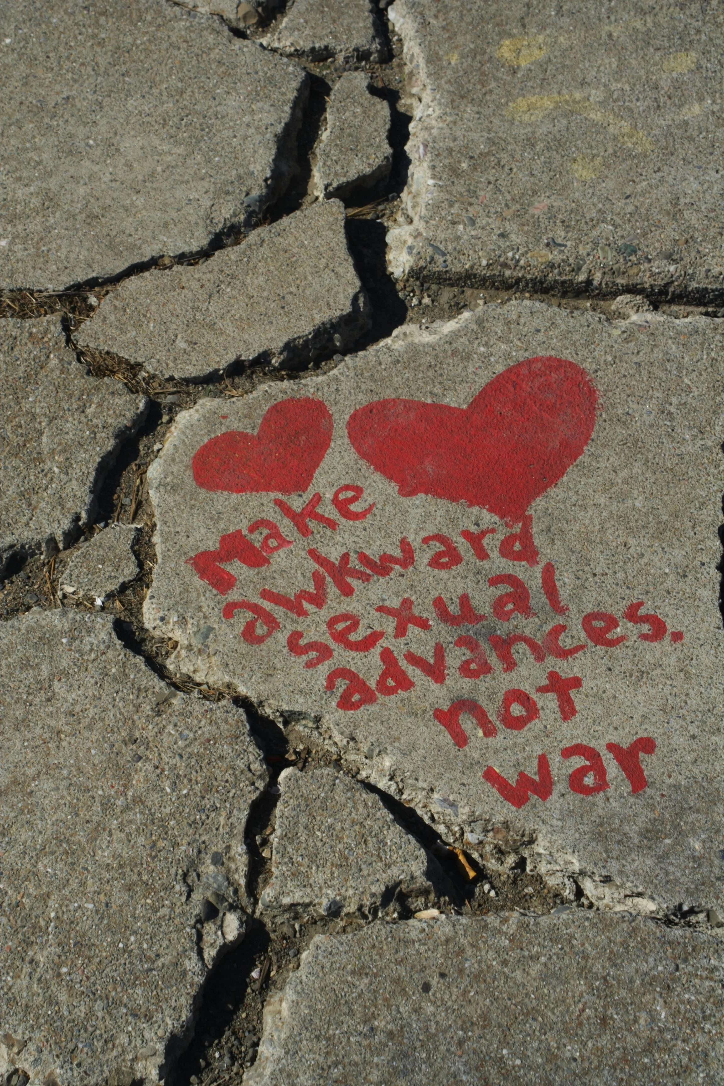 Concrete sidewalk with cracked pavement and a red stencil that says 'make awkward sexual advances. not war,' surrounded by two red hearts.