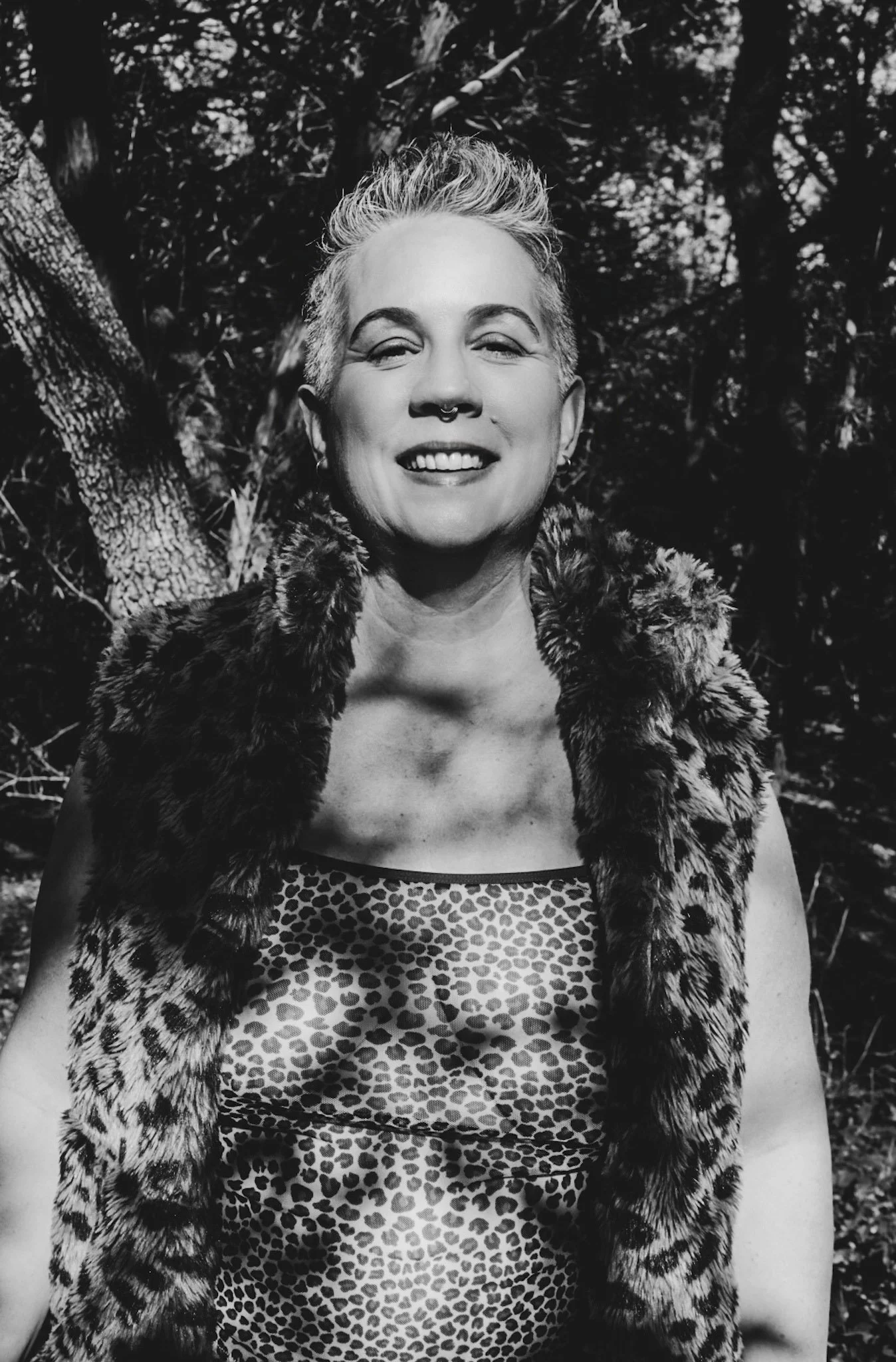 A smiling woman with short, light-colored hair, wearing a leopard-print tank and a fur patterned vest, standing outdoors in front of trees.