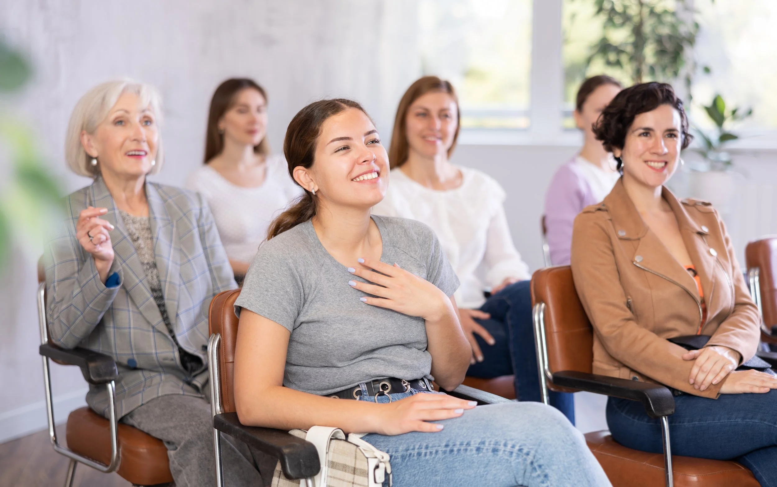 A group of women sitting and smiling in a bright room with large windows and green plants, attending a seminar or presentation.