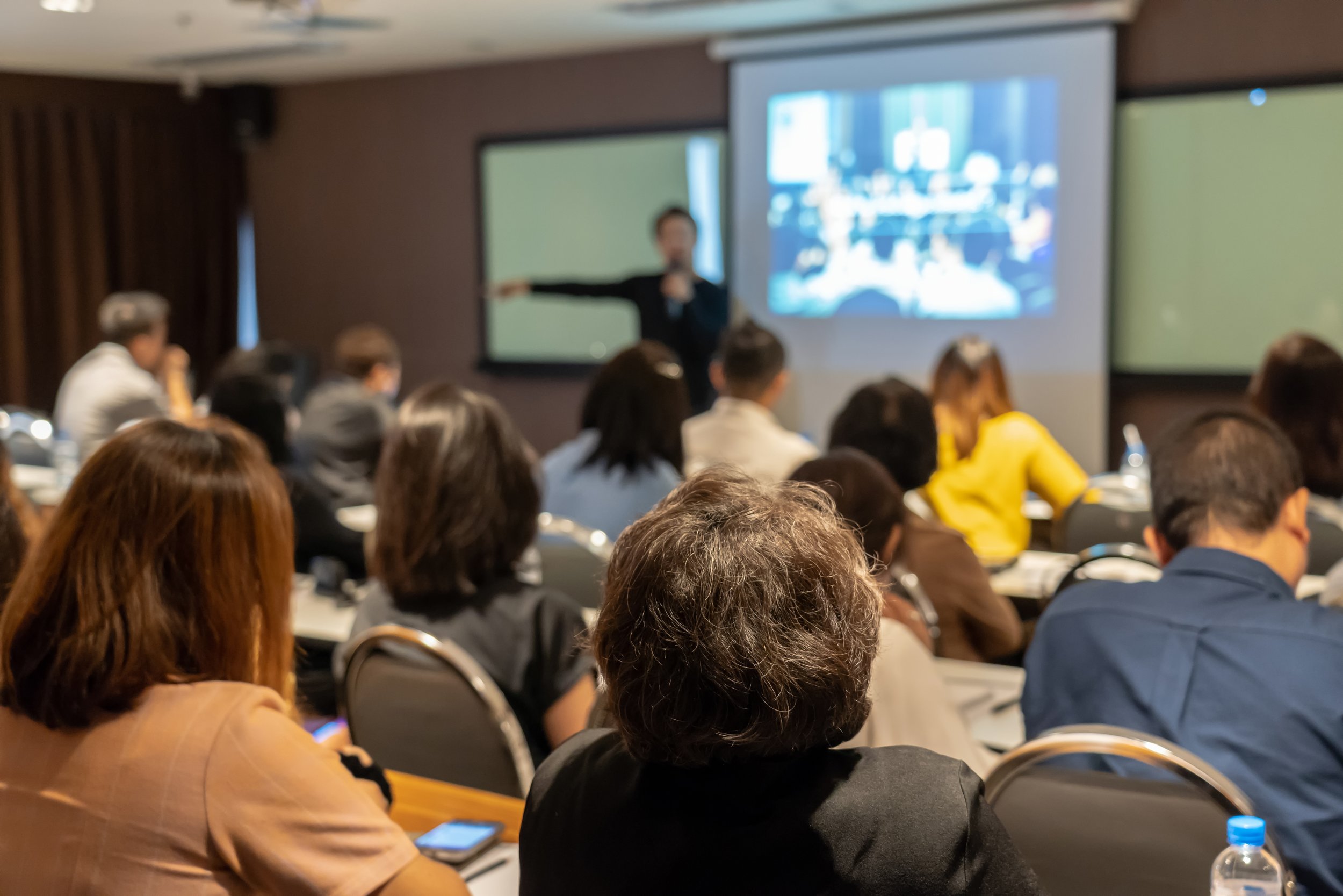 A group of people attending a presentation or seminar in a conference room, with a presenter pointing at a screen displaying a blurred image.